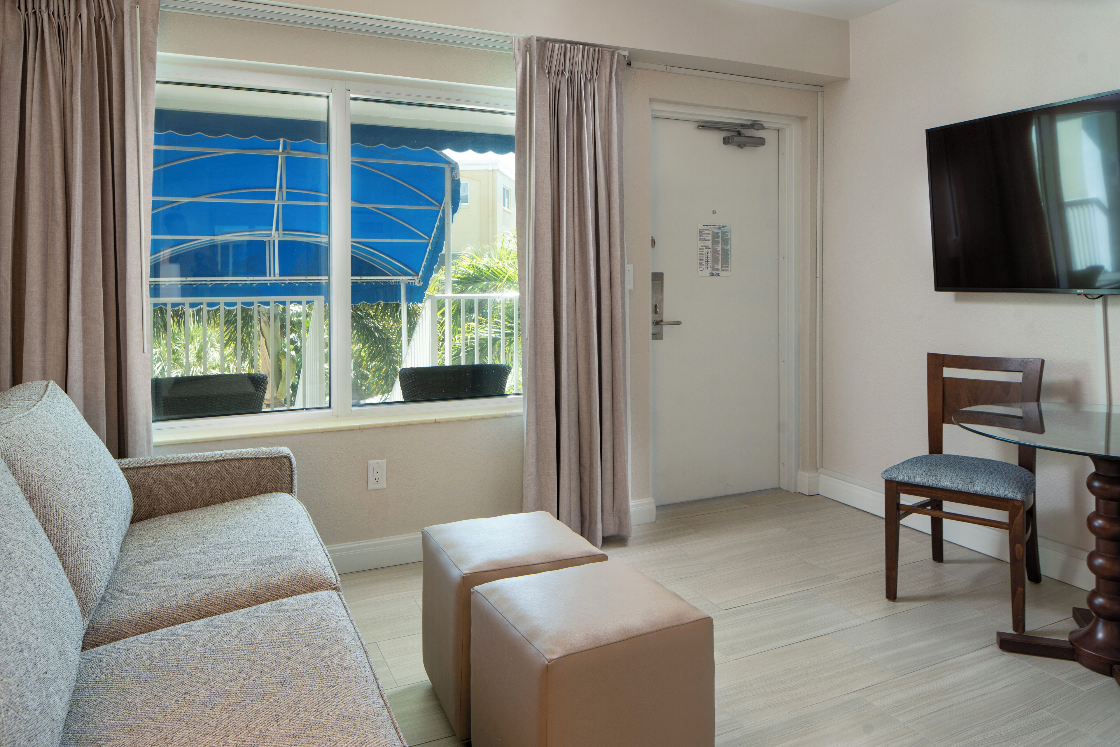 A living room with a sofa, ottoman, and a glass table with a chair. A television is mounted on the wall.