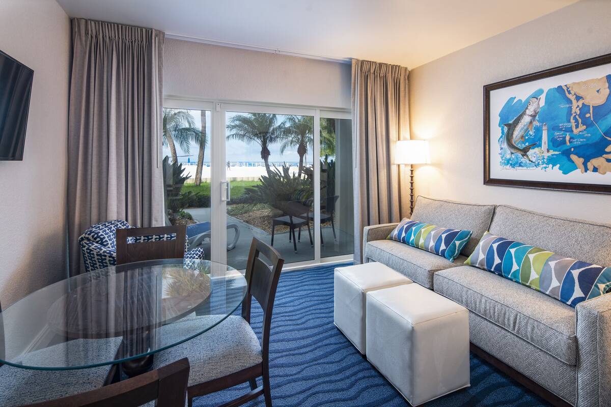 A hotel room with a glass dining table, a sofa with patterned cushions, a blue carpet, and a large framed map on the wall.
