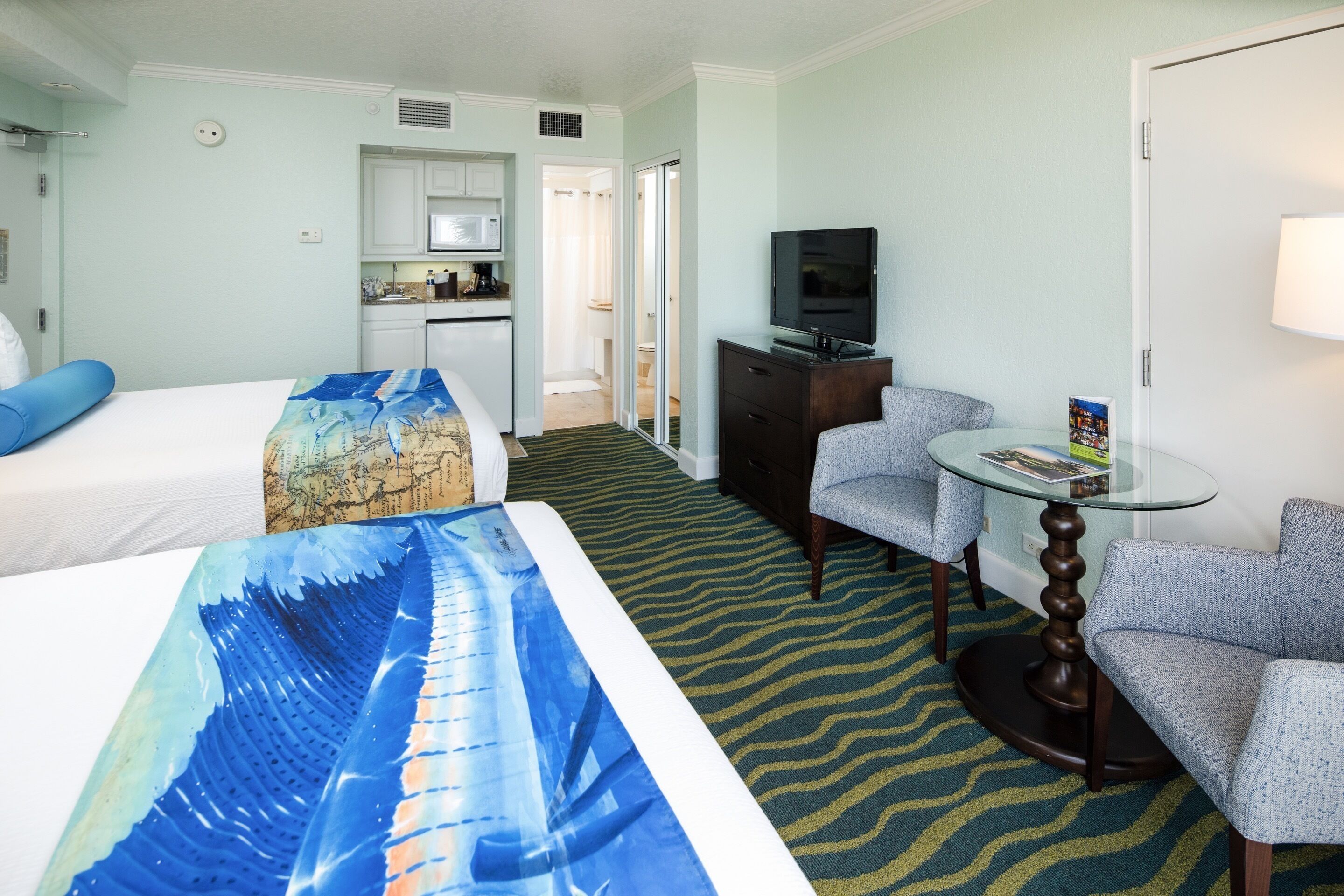 A hotel room with two beds, a small kitchenette, a TV, a round table, and a patterned carpet.