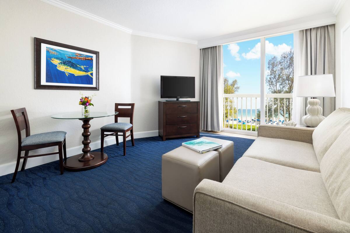 A hotel room with a balcony, a television, a sofa, and a dining area with chairs and a table.