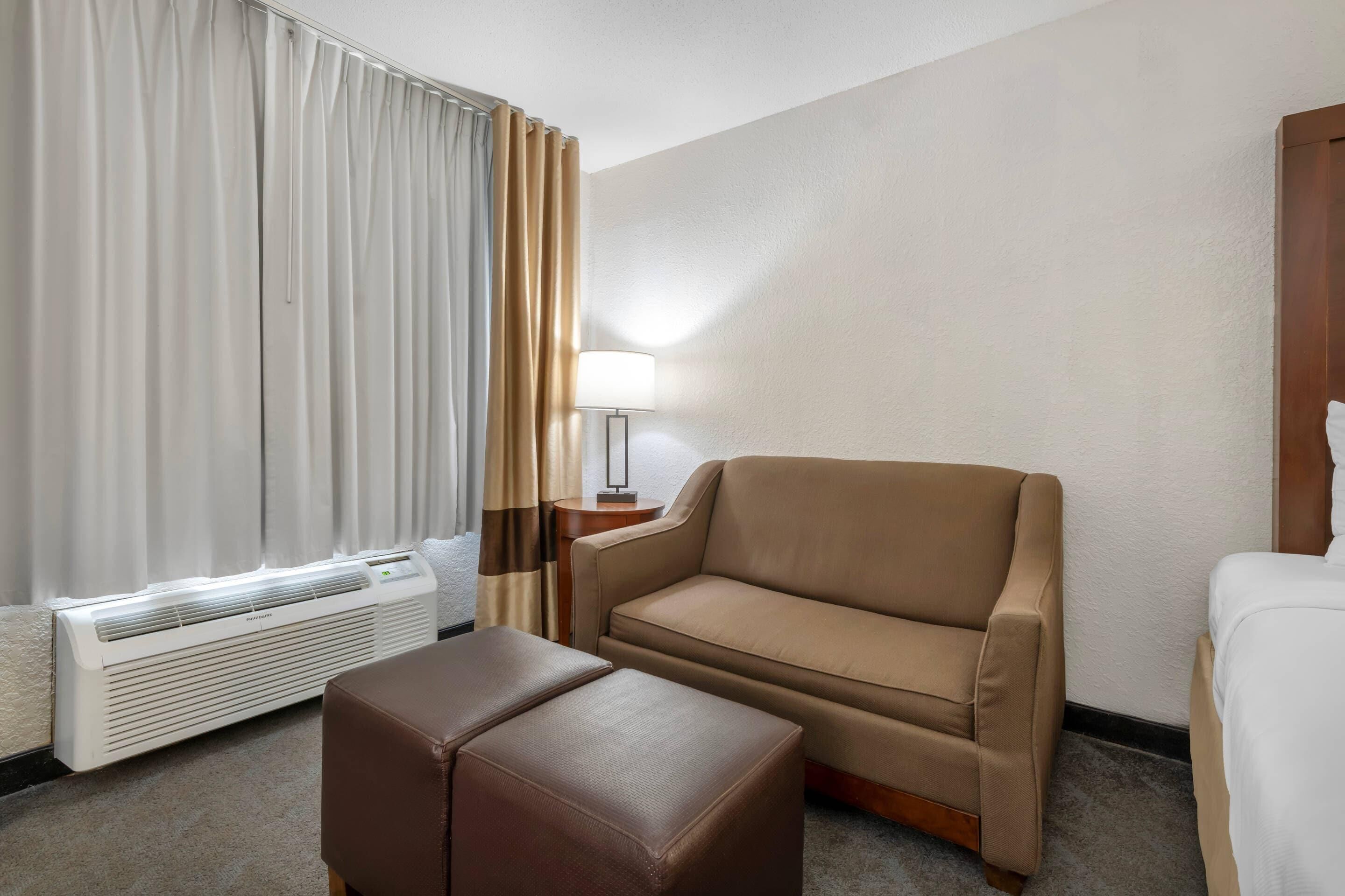 A hotel room with a beige sofa, a brown ottoman, a white bed, a lamp, and a window with curtains.