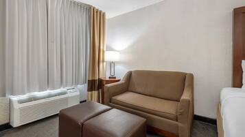 A hotel room with a beige sofa, a brown ottoman, a white bed, a lamp, and a window with curtains.