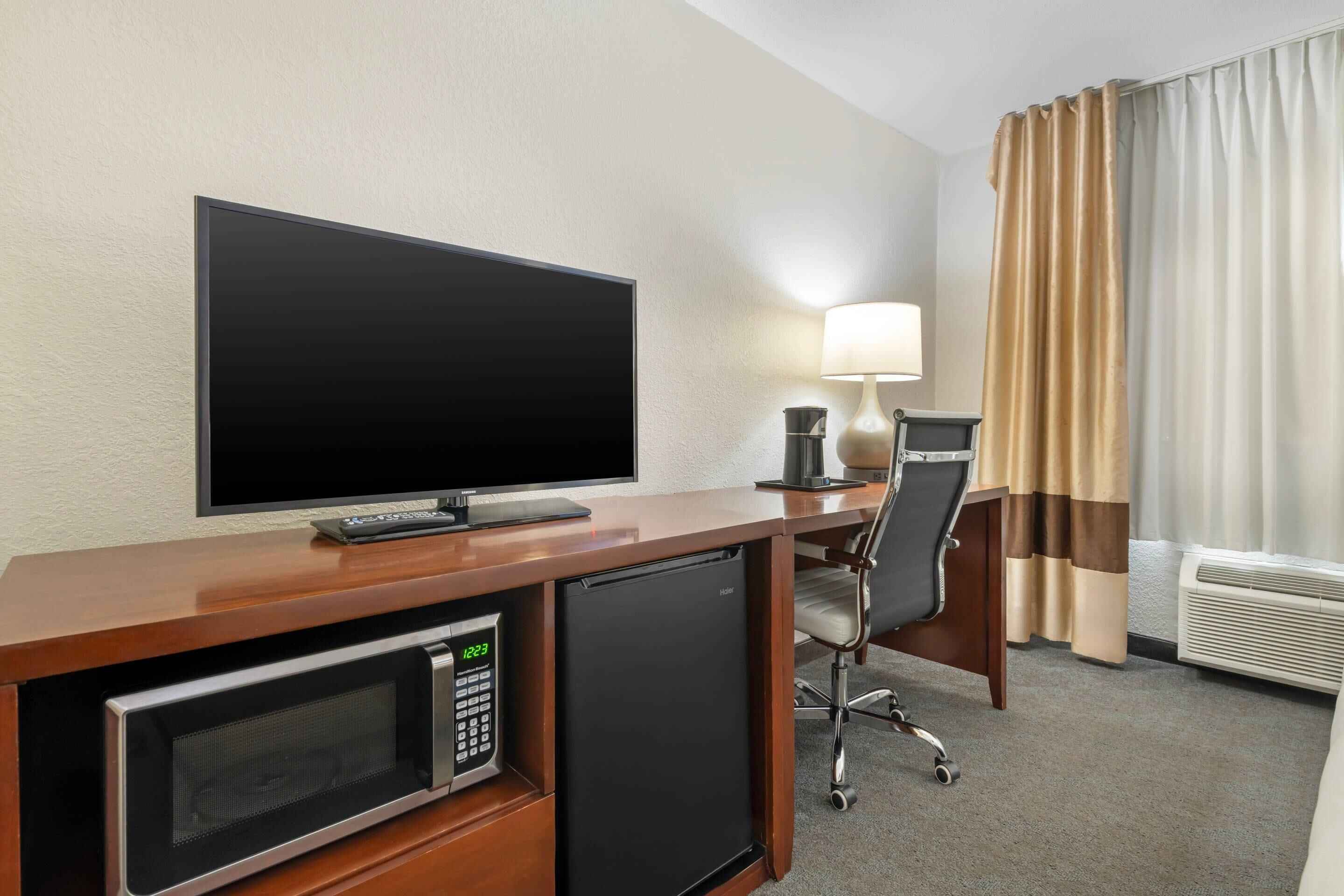 A hotel room with a flat-screen TV, a microwave, a mini-fridge, a desk with a lamp, a chair, and a window with curtains.