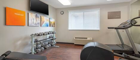 Un gimnasio con cinta de correr, mancuernas, banco, televisor montado en la pared y una ventana con persianas.