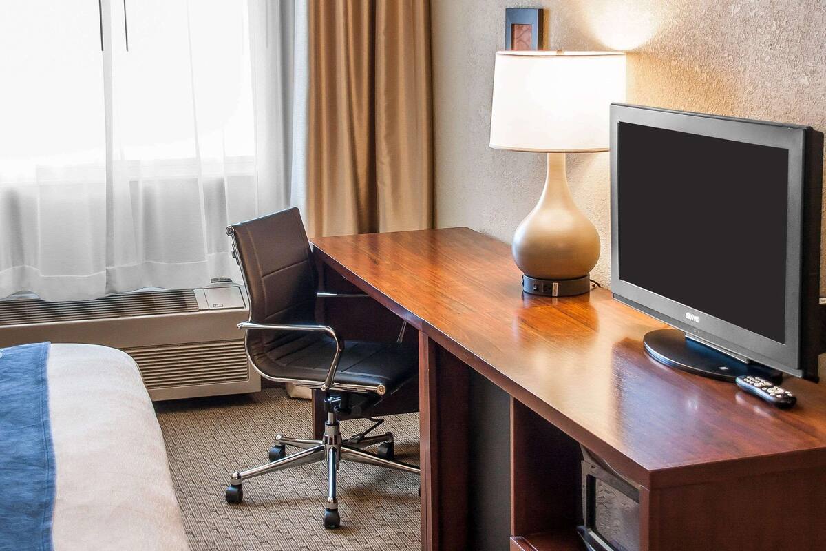 A hotel room with a wooden desk, a television, a chair, a lamp, and a bed.