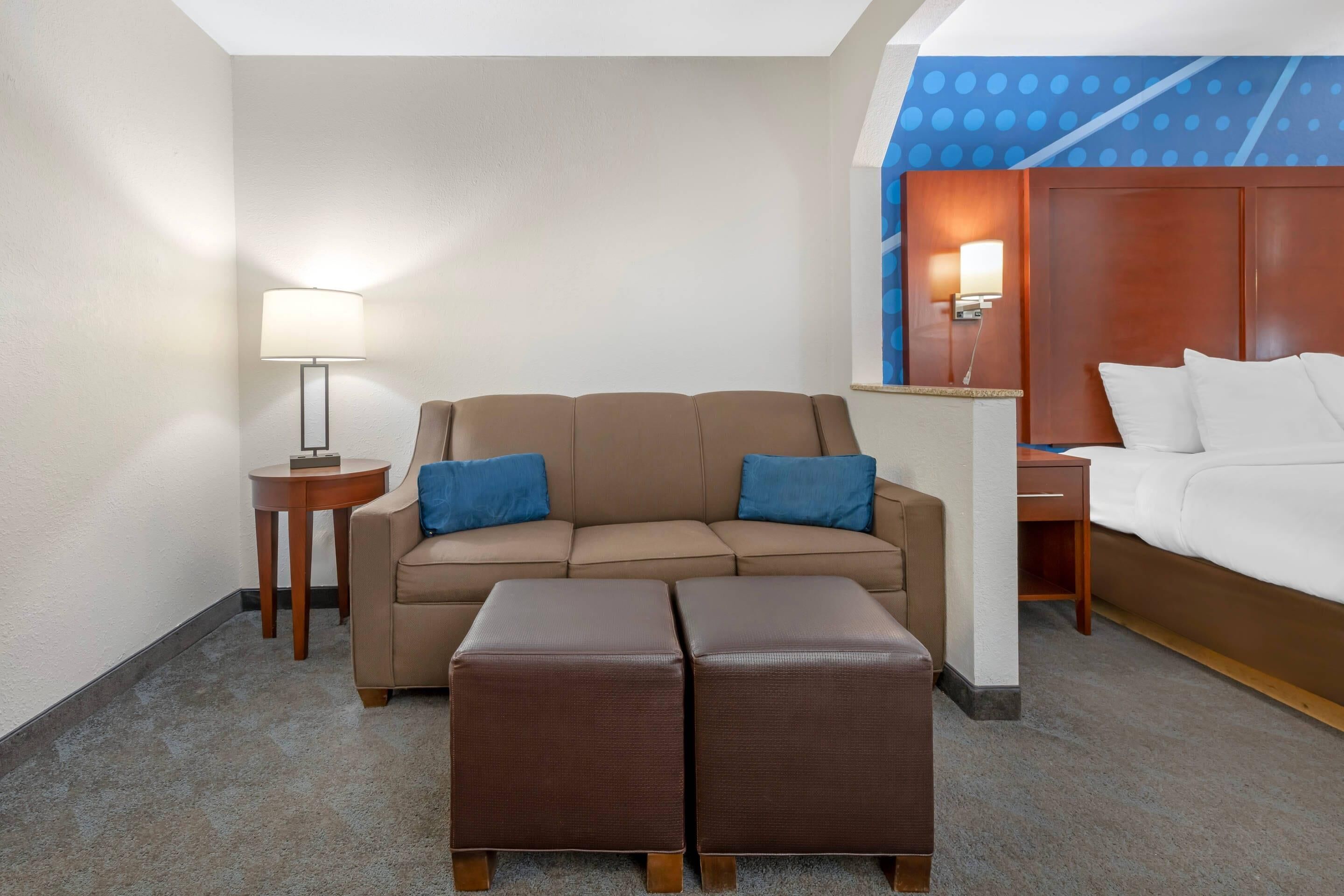 A hotel room with a sofa, two ottomans, a lamp, a bed, and a nightstand.