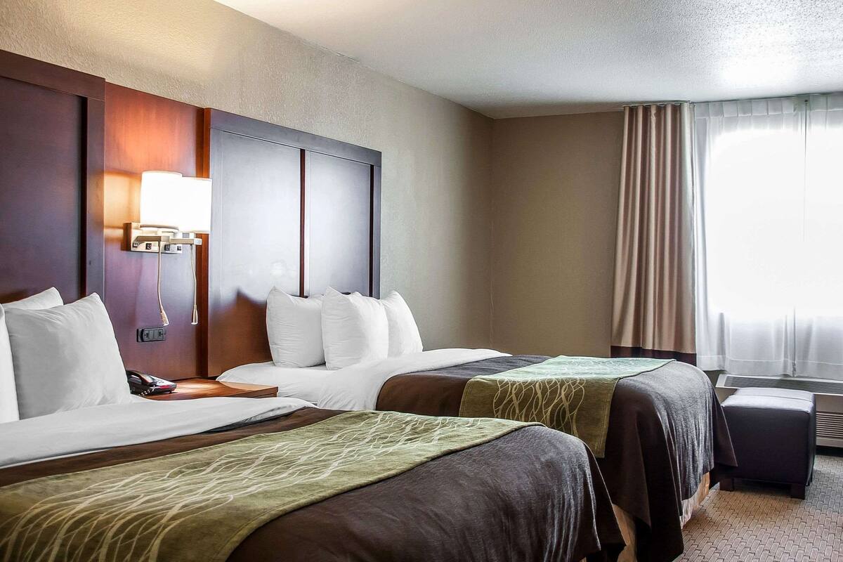 A hotel room with two beds, a bedside table with a lamp, an air conditioning unit, and a window with curtains.