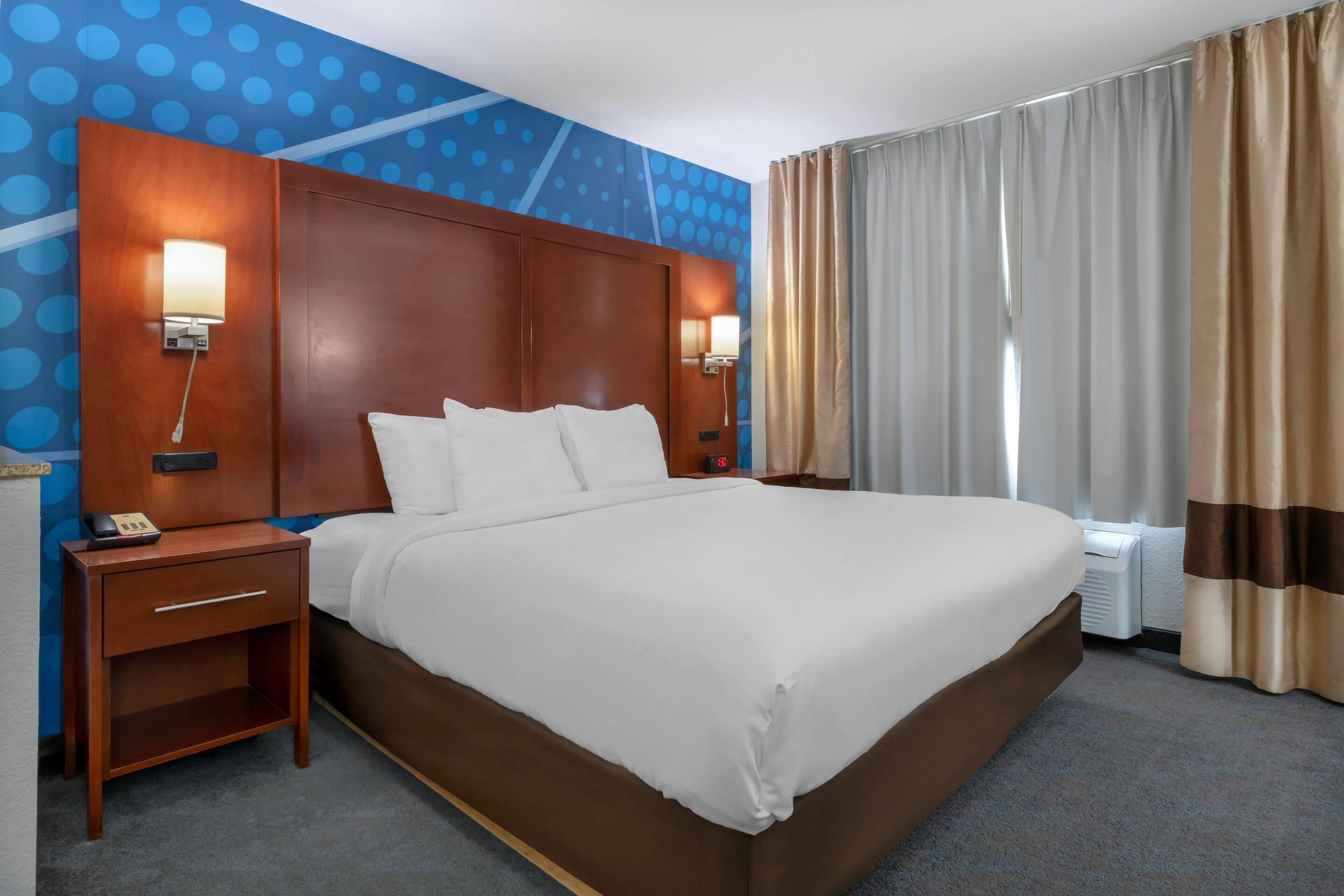 A hotel room with a large bed, two bedside tables with lamps, a nightstand, a wall-mounted phone, and a window with curtains.
