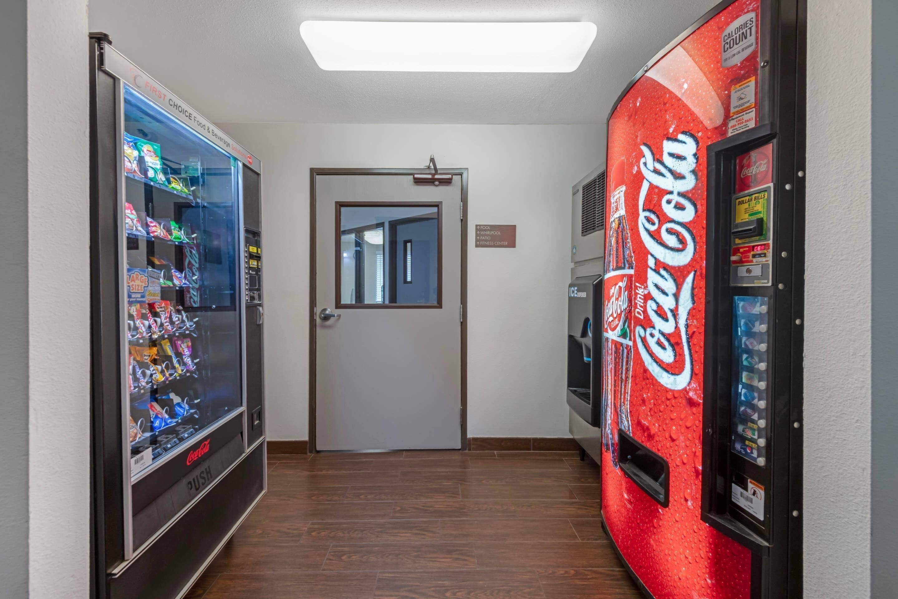 A hallway with vending machines, including a Coca-Cola branded one, and a door leading to another room.