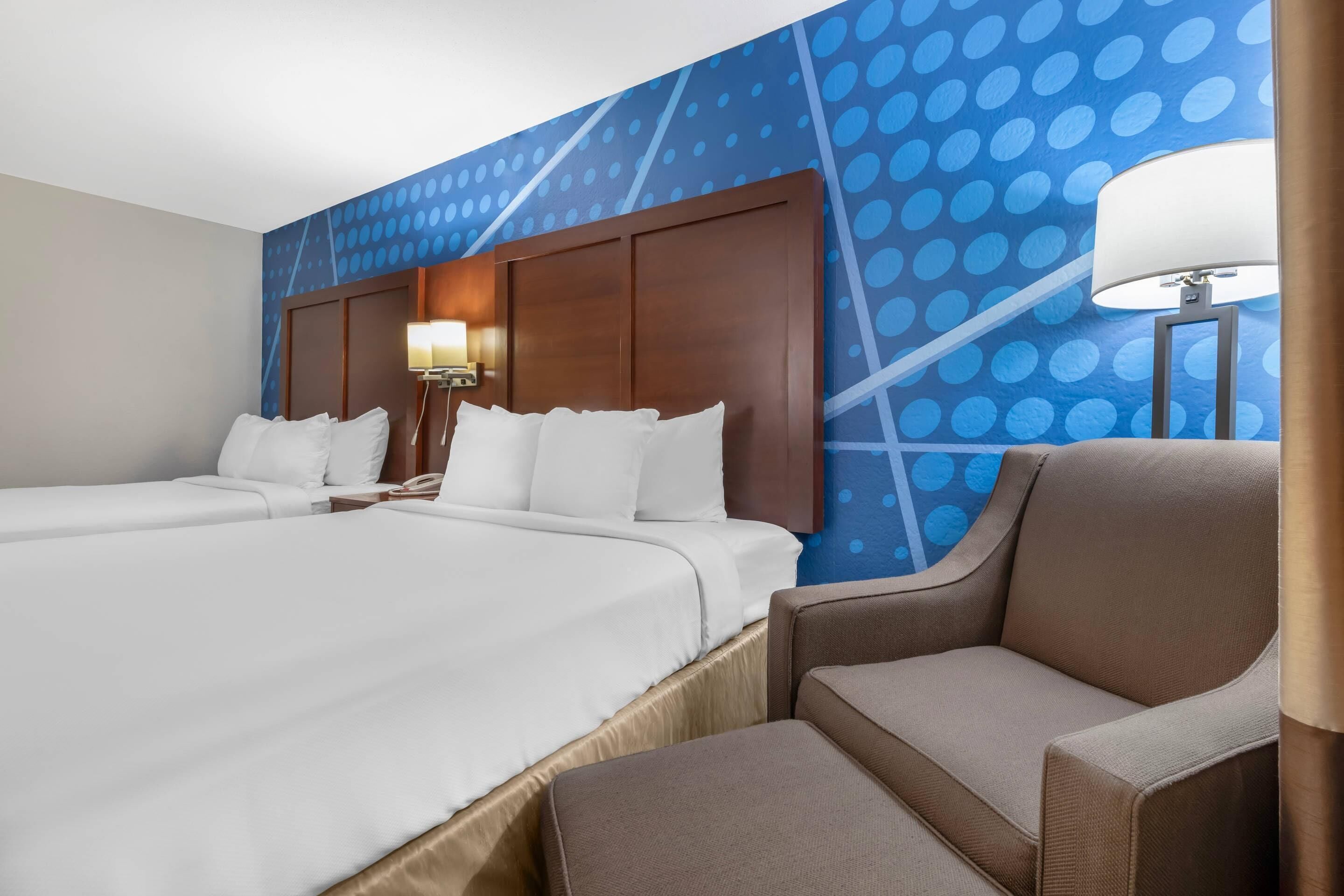 A hotel room with two beds, a nightstand, a lamp, and a chair.