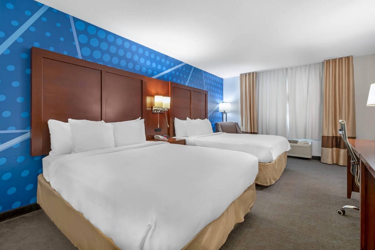 A hotel room with two beds, a desk, a lamp, and a large window with curtains.
