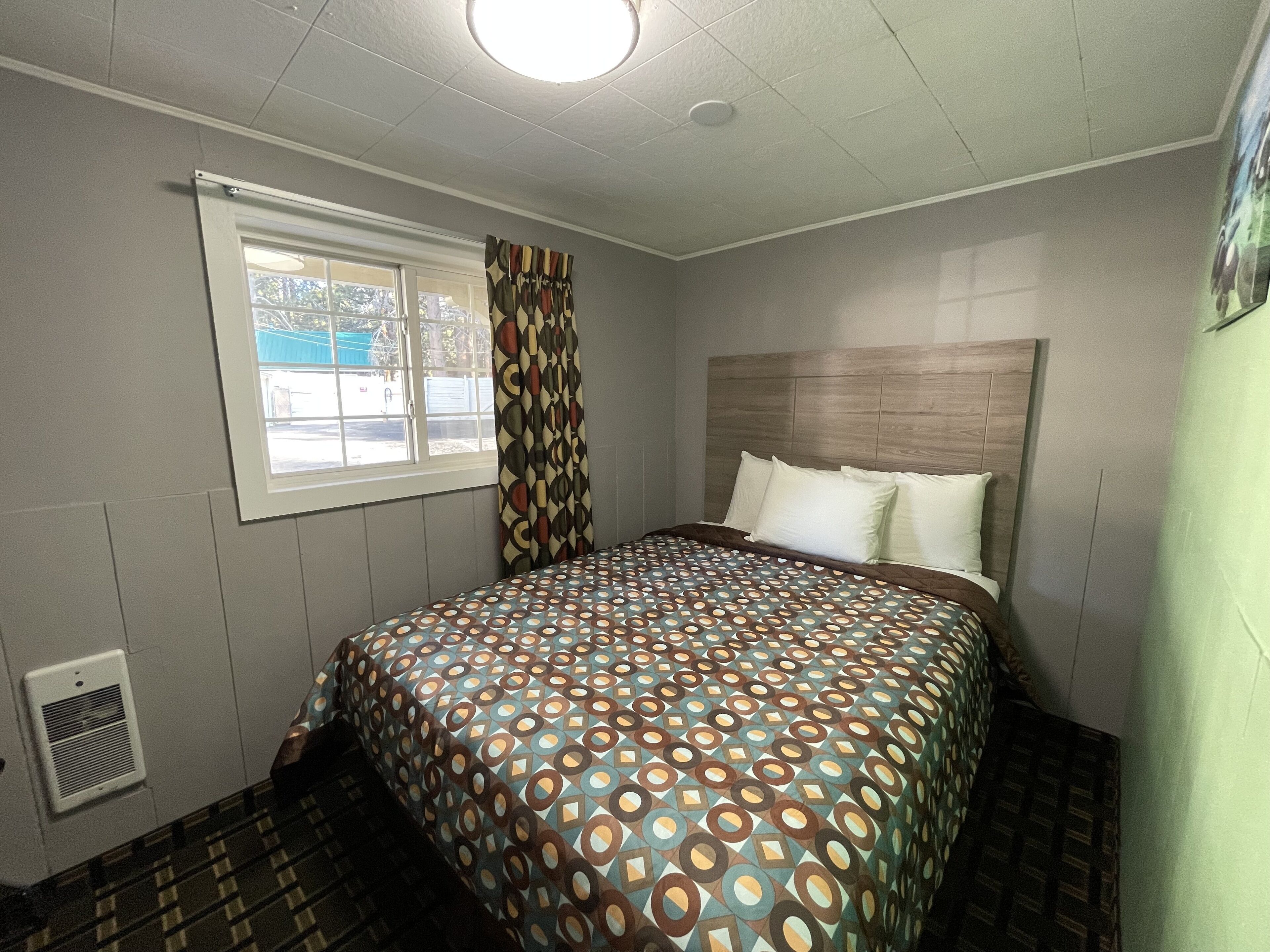 A bedroom with a patterned bedspread, a window with curtains, a radiator, and a headboard.
