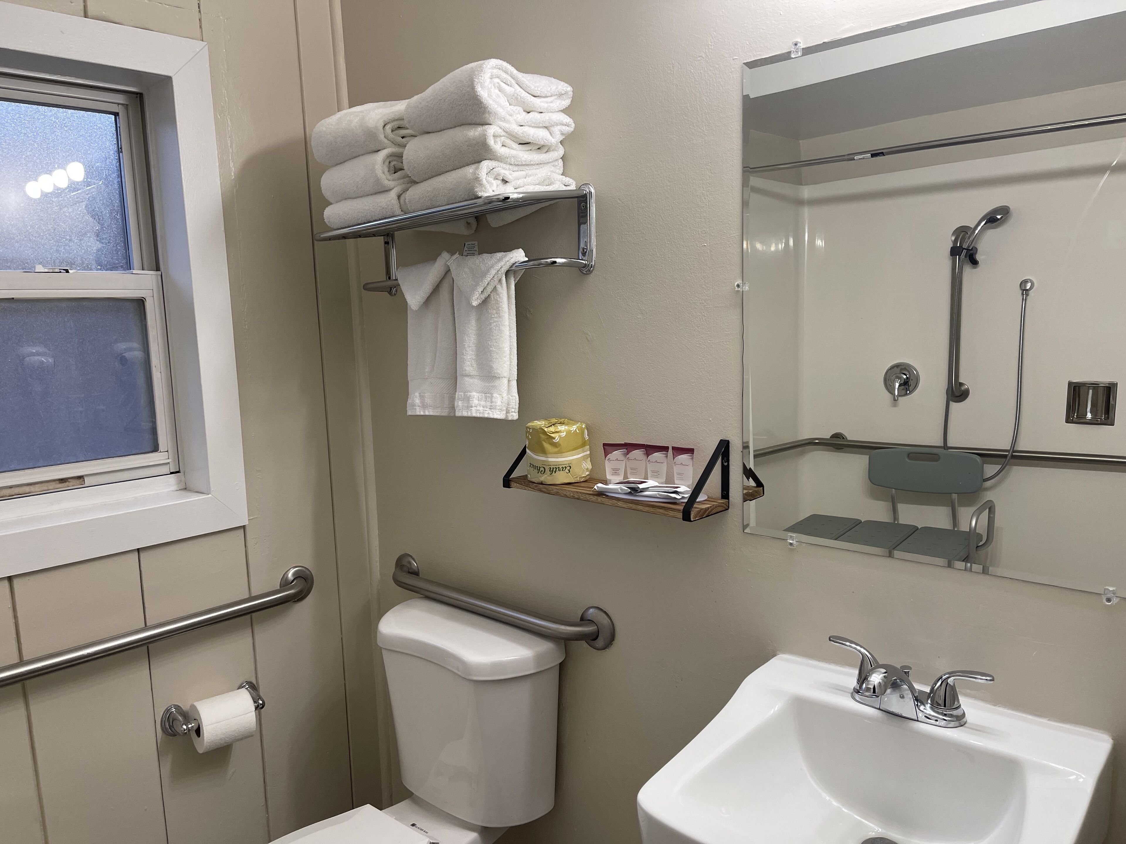 Baño con inodoro, lavamanos y zona de ducha. Hay una ventana, un perchero y un estante con artículos de aseo.