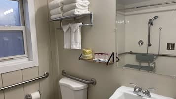 Baño con inodoro, lavamanos y zona de ducha. Hay una ventana, un perchero y un estante con artículos de aseo.