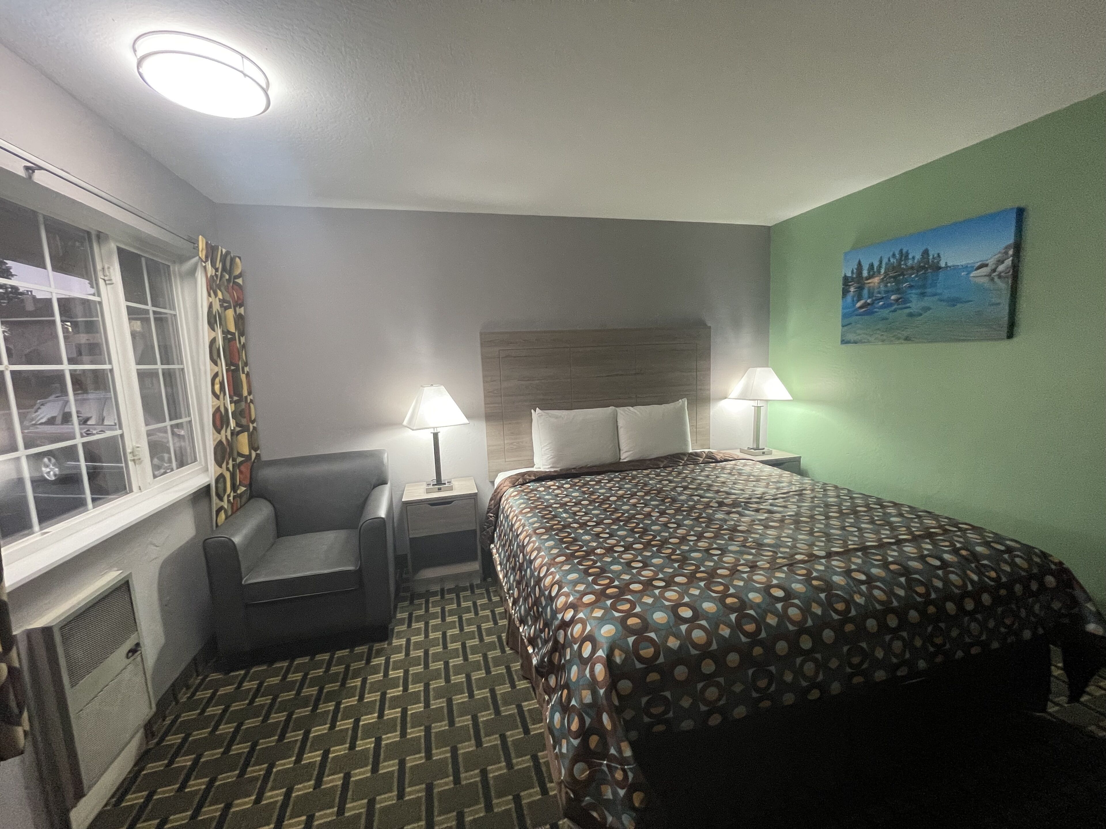 A hotel room with a bed, two bedside lamps, a chair, a window with curtains, and a painting on the wall.