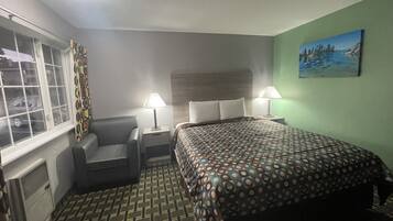 A hotel room with a bed, two bedside lamps, a chair, a window with curtains, and a painting on the wall.
