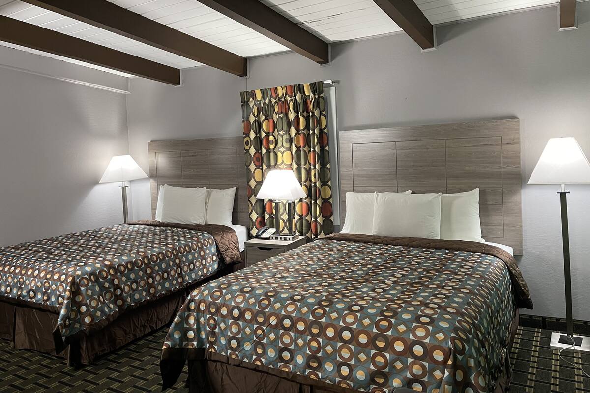 A hotel room with two beds, each with patterned bedding, a headboard, bedside lamps, and a curtain with a geometric pattern.
