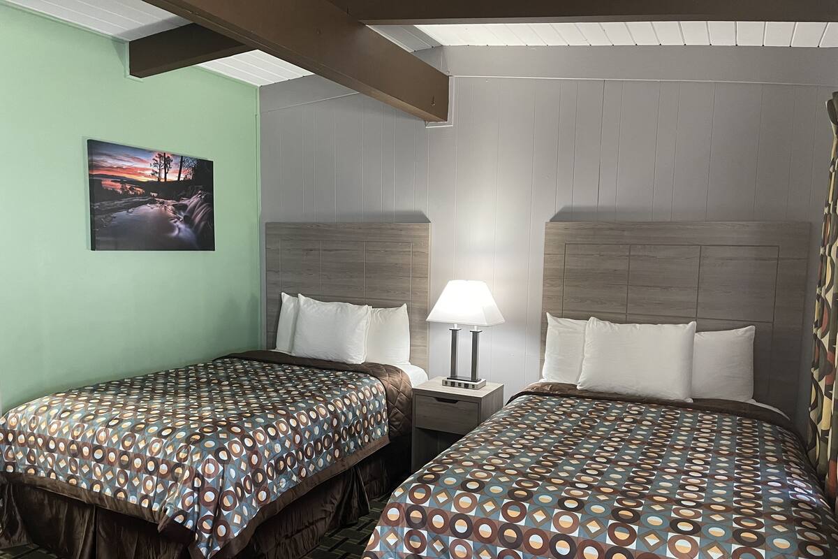 A hotel room with two beds, a nightstand with a lamp, a painting on the wall, and a patterned bedspread.