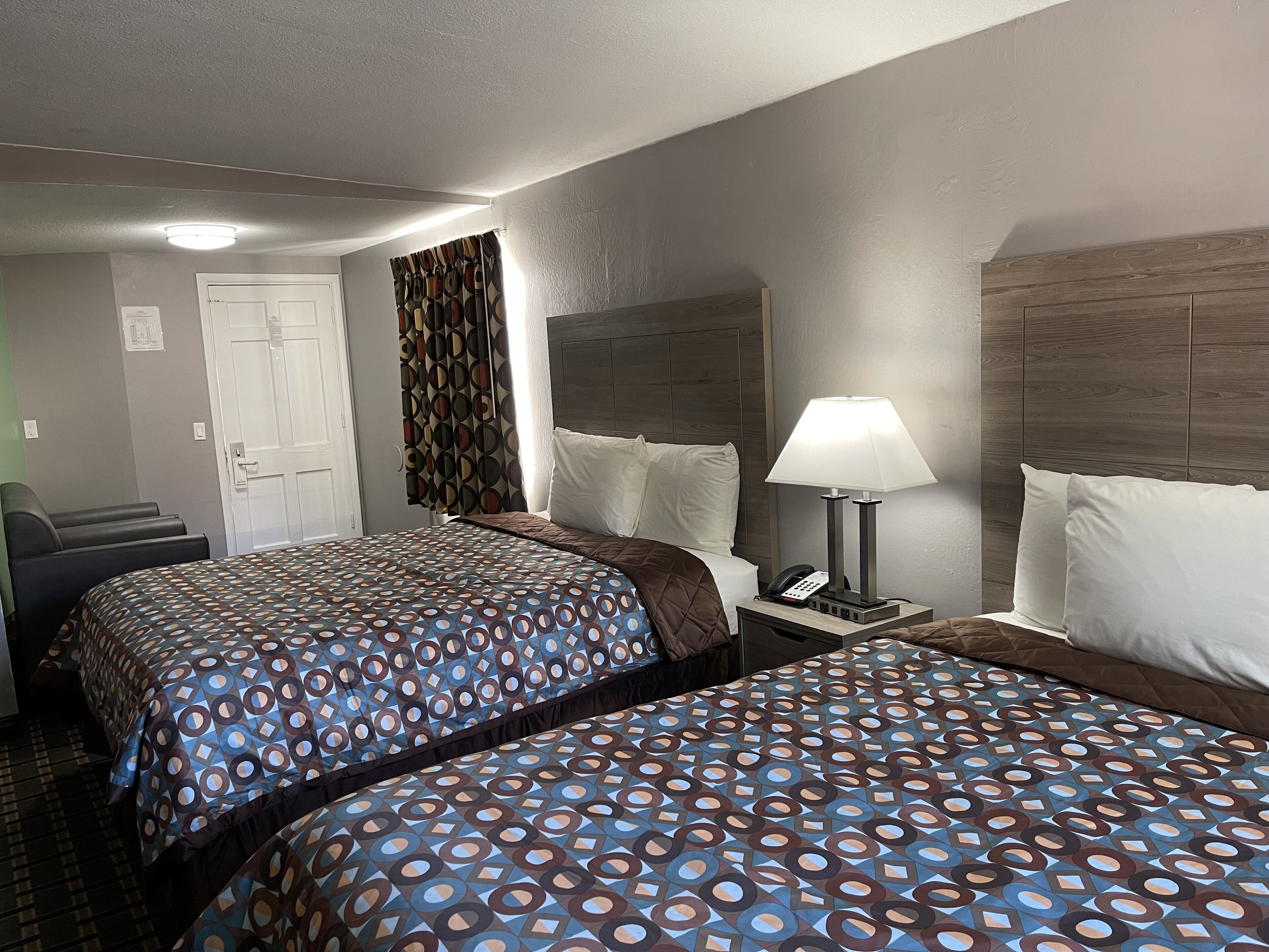 A hotel room with two beds, a bedside table with a lamp, a telephone, and a door.
