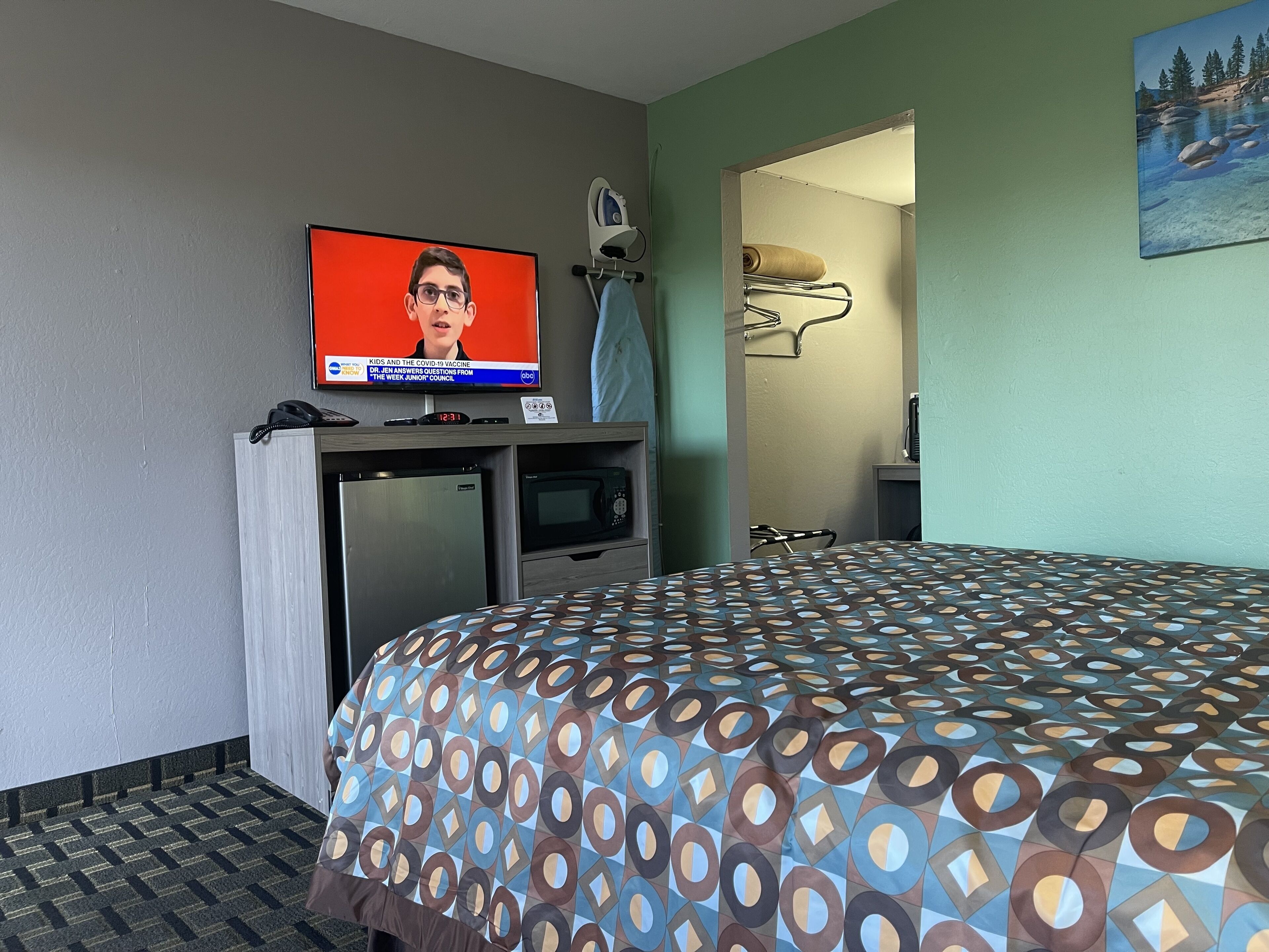 A hotel room with a bed, a television displaying a news program, a desk with a computer and a phone, and a microwave.