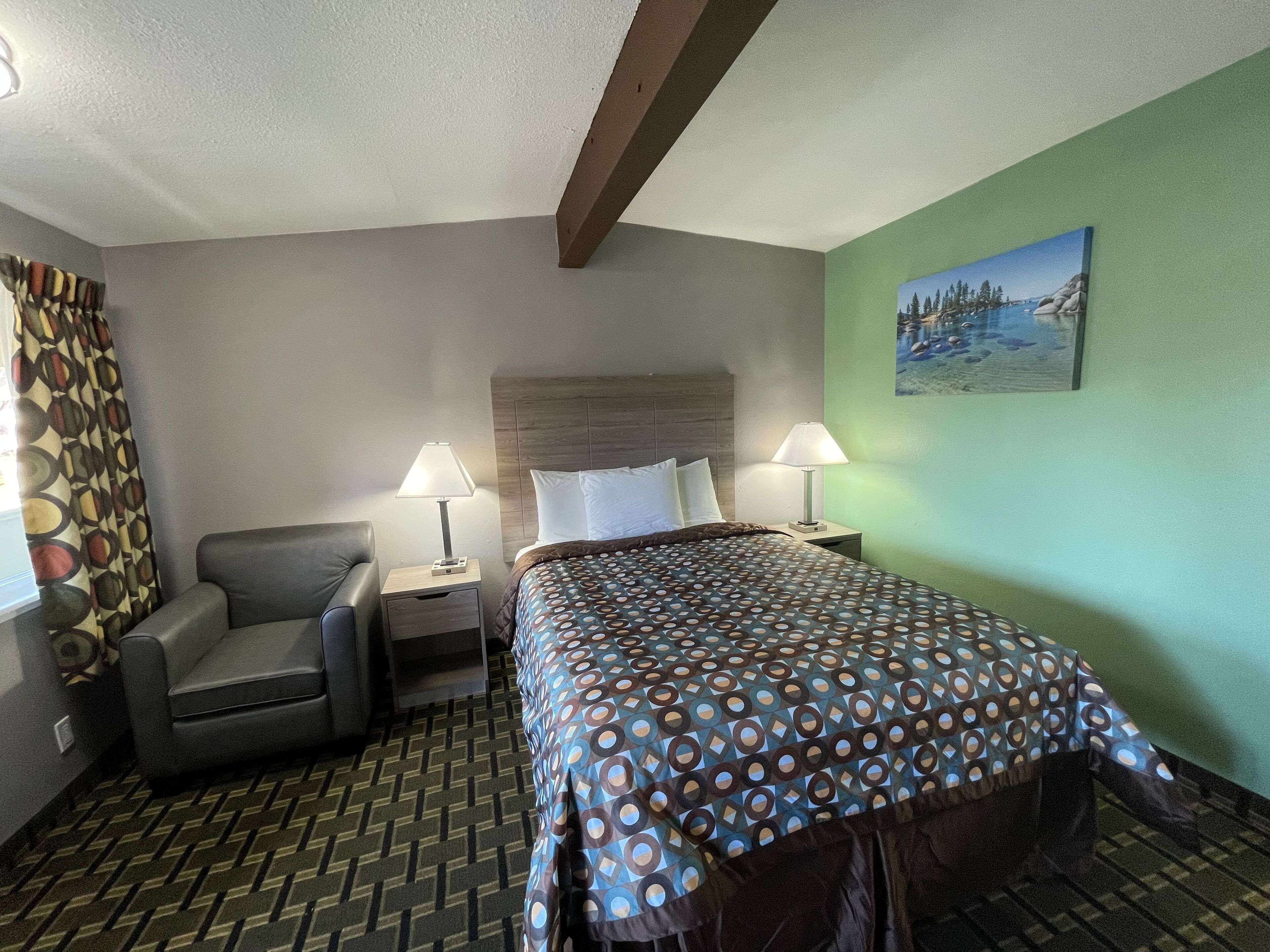 A hotel room with a bed, two bedside tables with lamps, a chair, a patterned carpet, and a painting on the wall.
