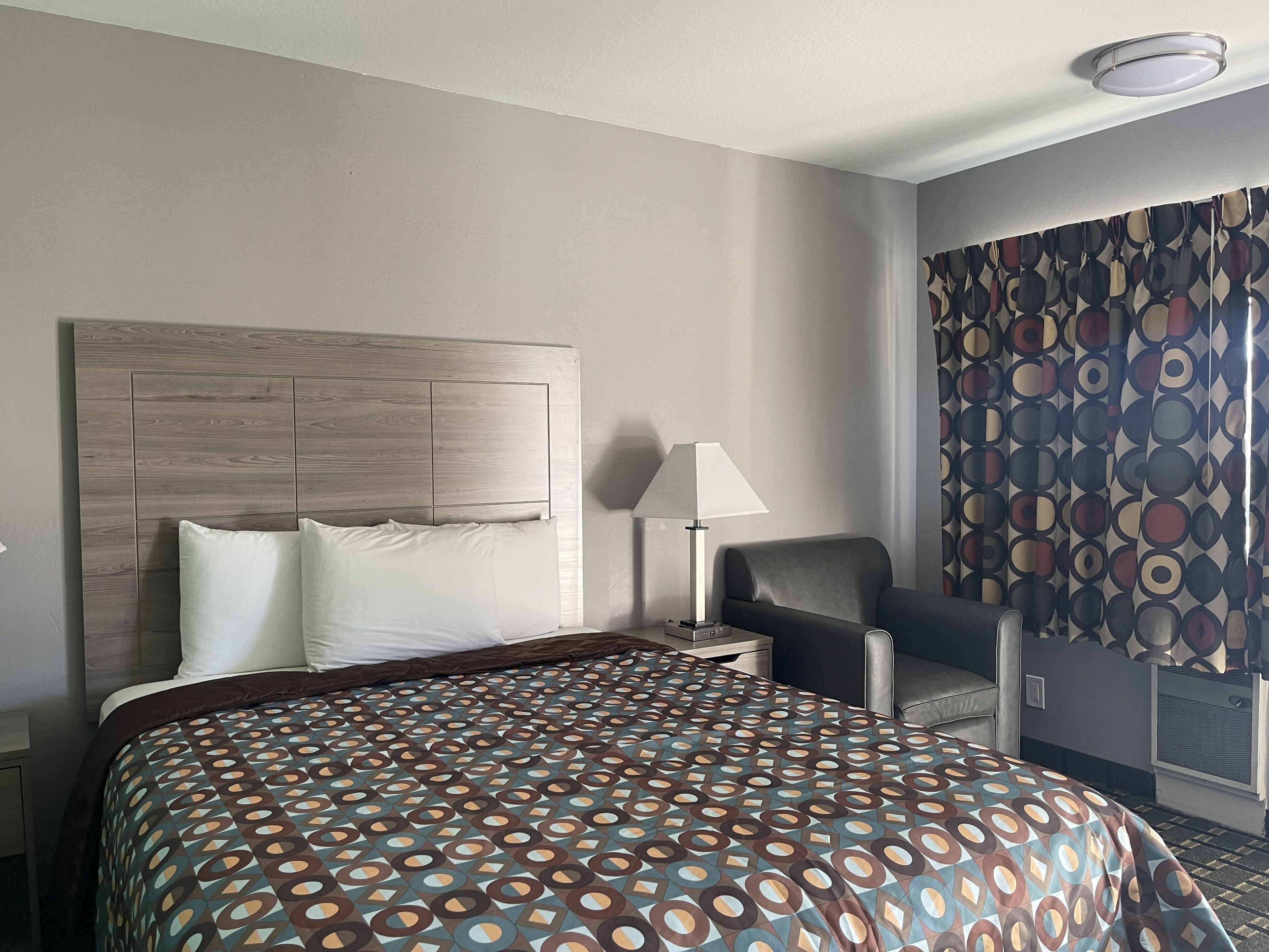 Een hotelkamer met een groot bed, een nachtkastje, een bureau, een stoel en een raam met patroon gordijnen.