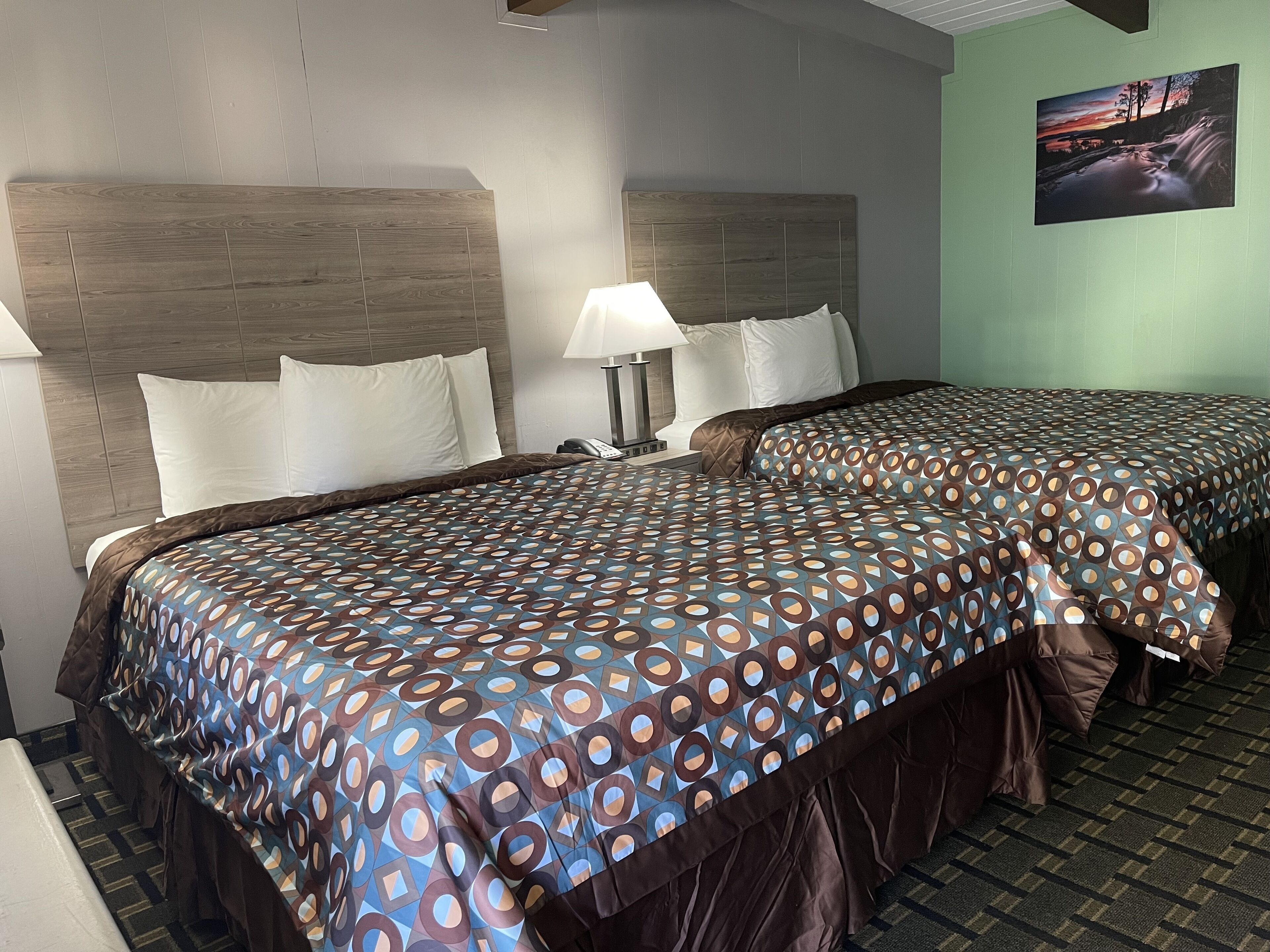 Habitación de hotel con dos camas, cabecero, mesita de noche, lámpara y un cuadro en la pared.