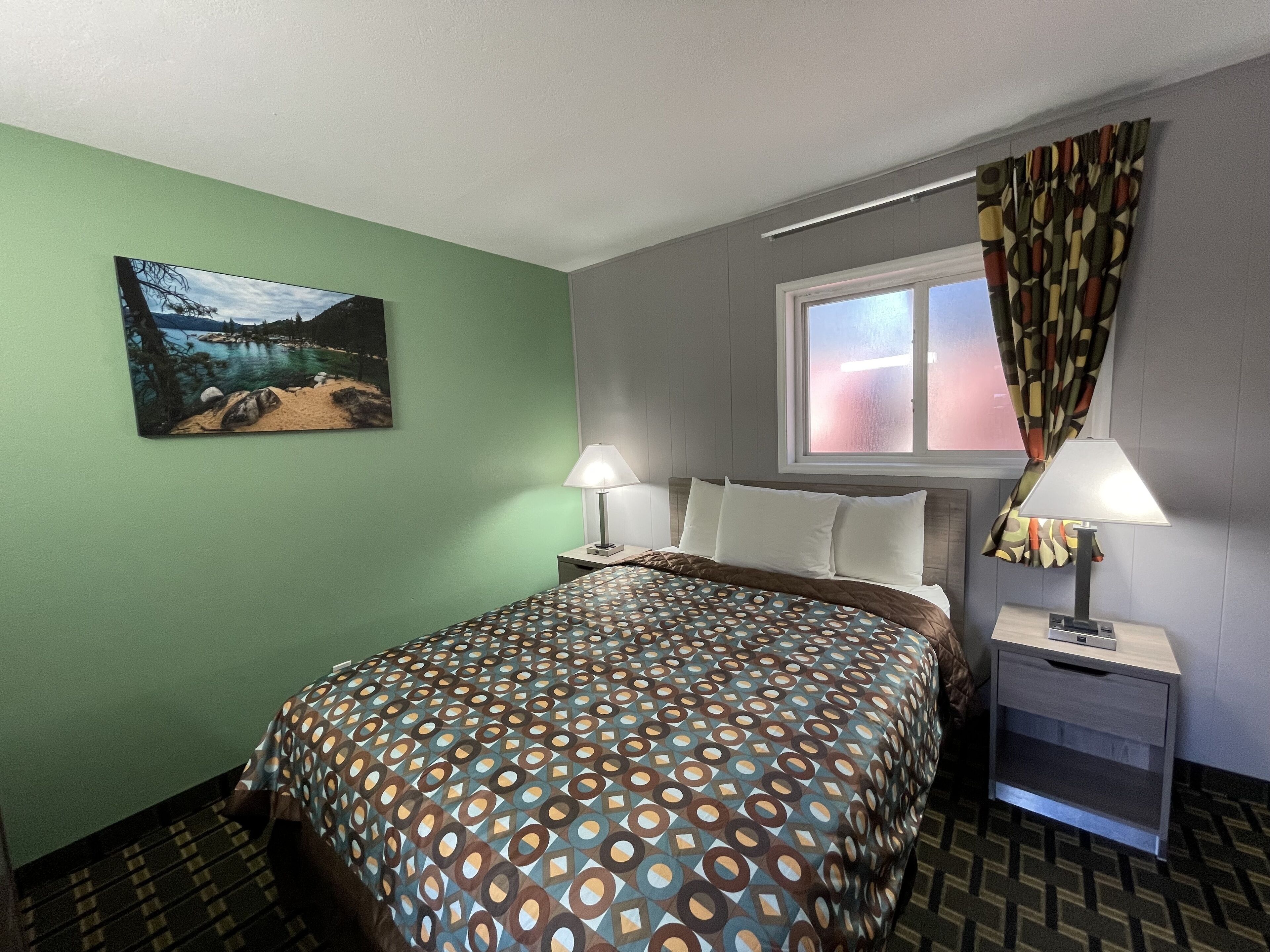 Habitación de hotel con cama, dos mesitas de noche, una ventana con cortinas, un cuadro en la pared y una colcha estampada.