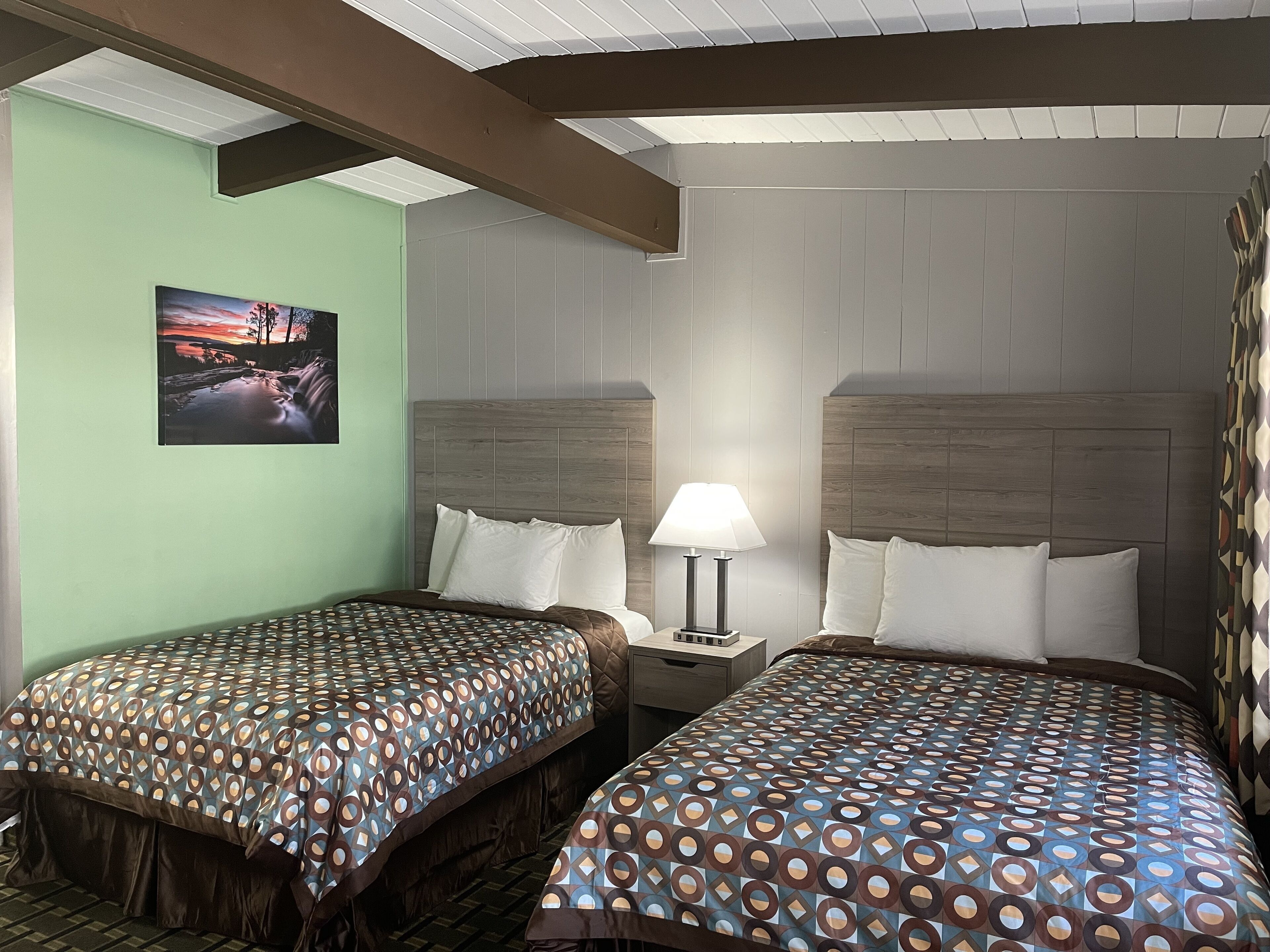 A hotel room with two single beds, a nightstand with a lamp, a painting on the wall, and a patterned bedspread.