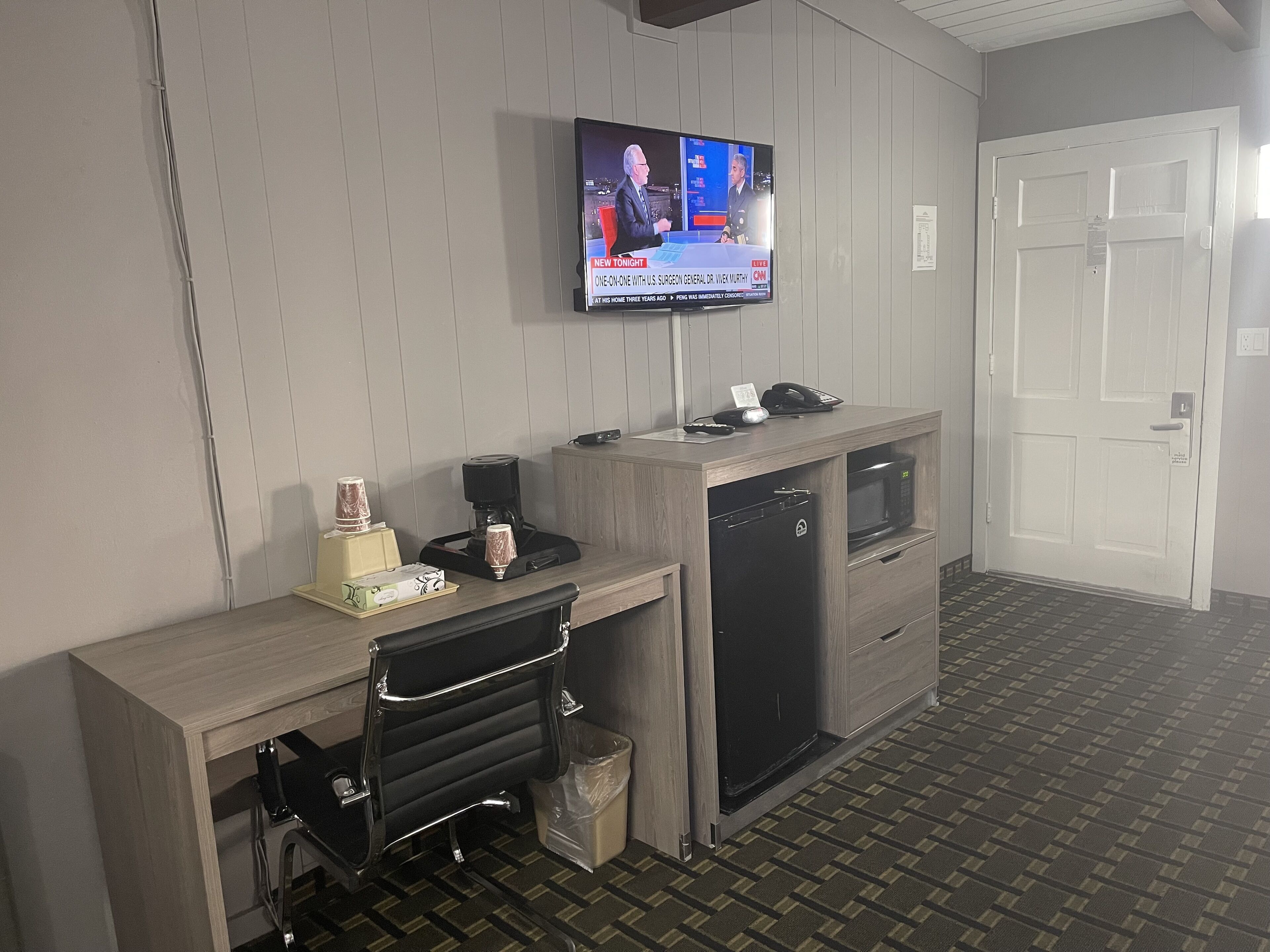 A hotel room with a wooden desk, a television mounted on the wall, a microwave, and a refrigerator.