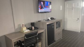 A hotel room with a wooden desk, a television mounted on the wall, a microwave, and a refrigerator.