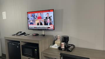 A hotel room with a television displaying a news broadcast, a desk with a phone, a microwave, and a coffee maker.