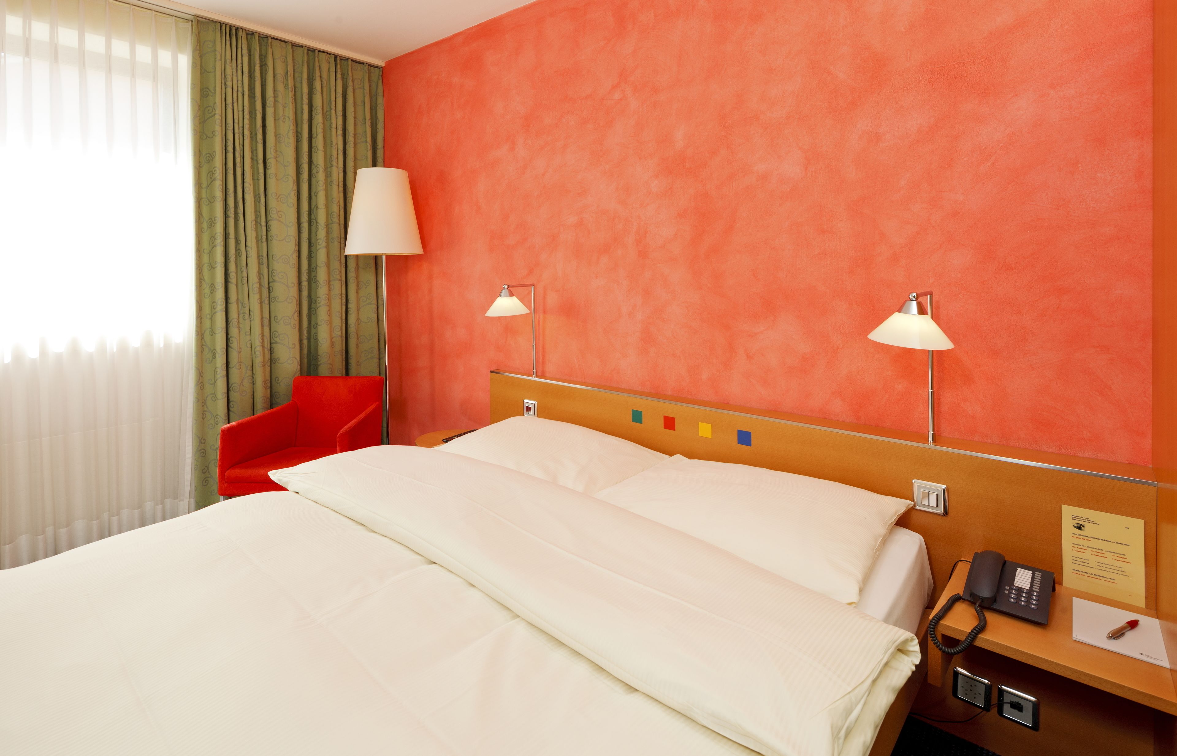 A hotel room with a bed, a red chair, two bedside lamps, a telephone, and a wall-mounted control panel.