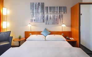 A hotel room with a large bed, two bedside lamps, a small table with a plant, a blue pillow, and a phone on the nightstand.