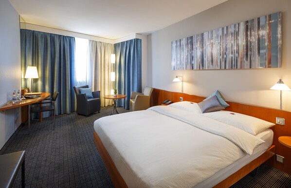 A hotel room with a large bed, a desk, a chair, a small table, and a lamp.