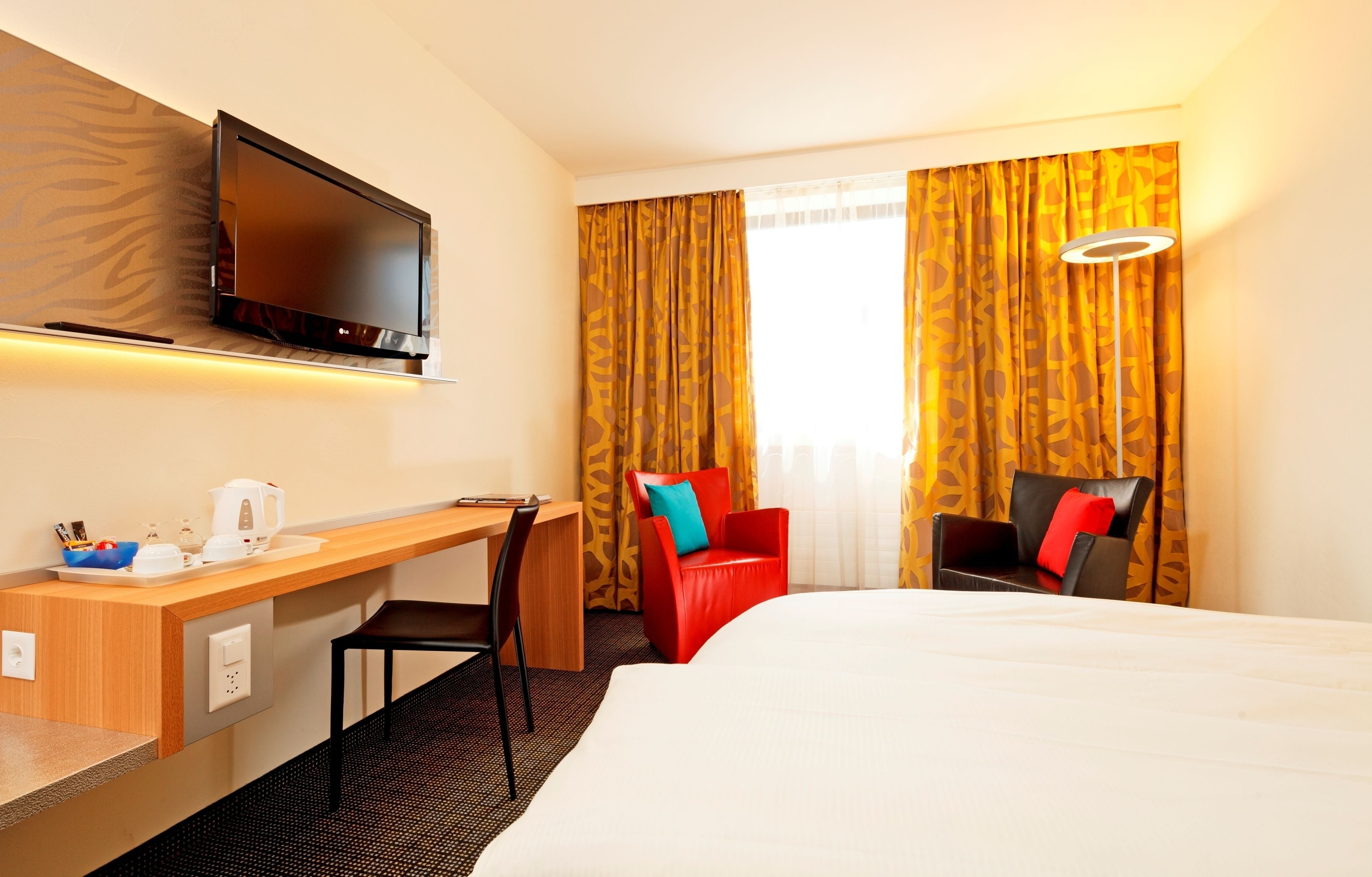 Ein Hotelzimmer mit Bett, Schreibtisch mit Stuhl, Fernseher und einem Fenster mit Vorhängen.