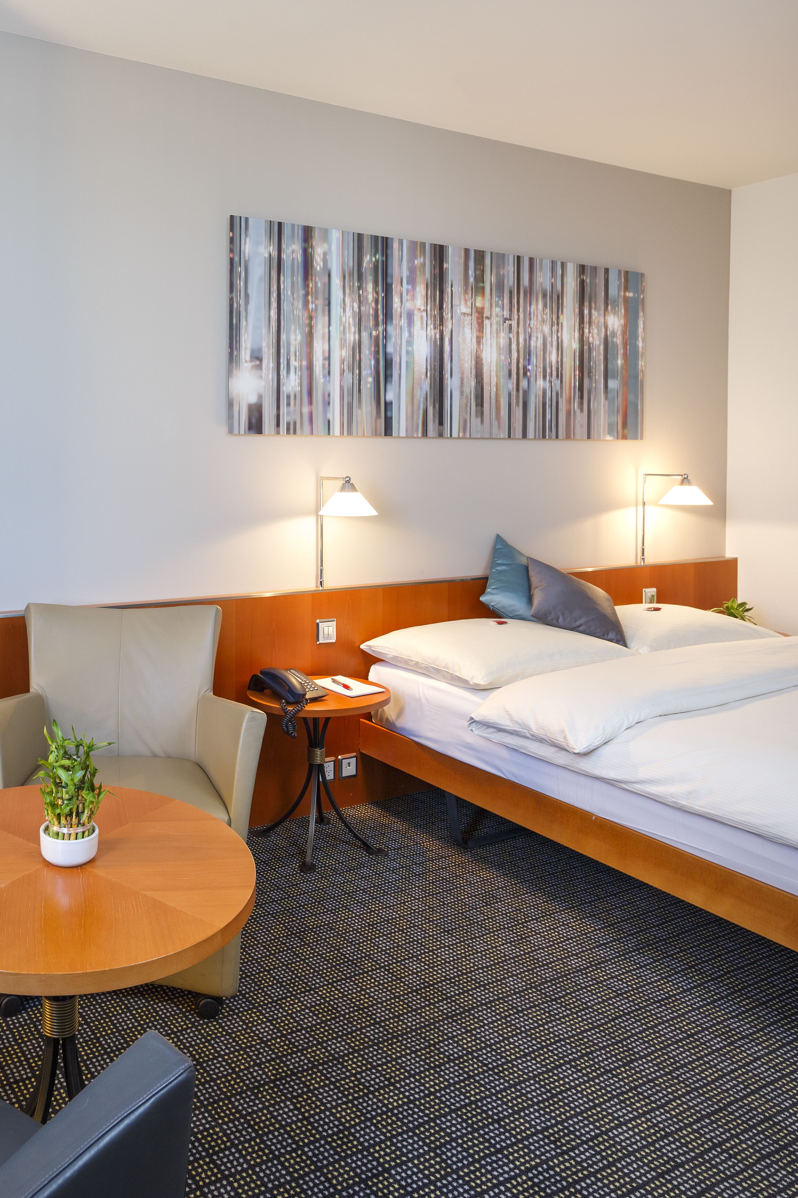 A hotel room with a bed, two bedside tables, a desk, a chair, a potted plant, and a wall-mounted artwork.