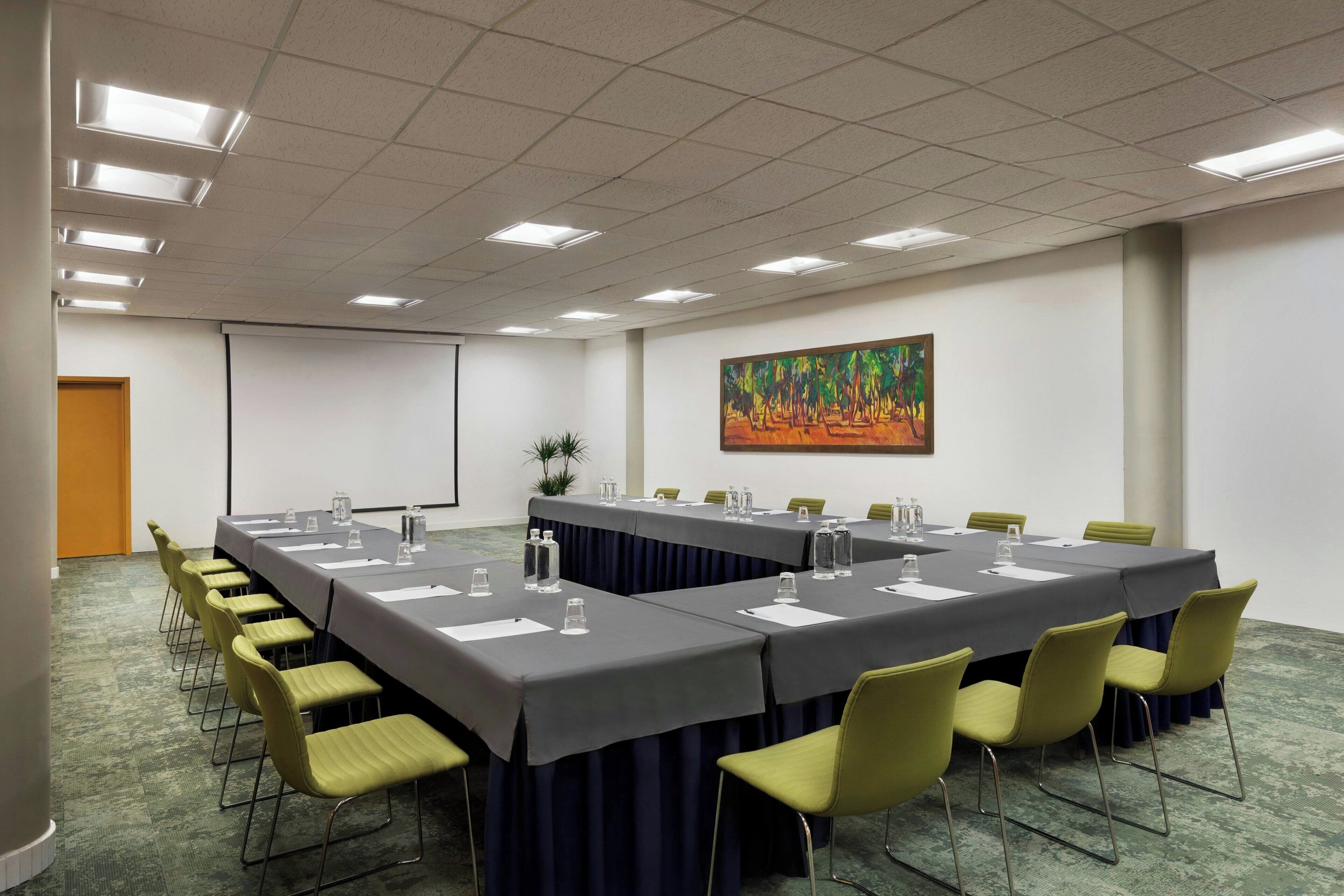 Un salón de conferencias con una larga mesa, sillas, una pantalla de proyector y un cuadro en la pared.
