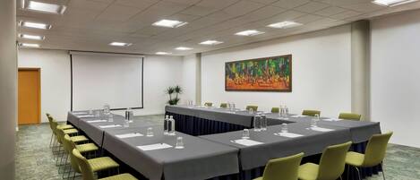 Un salón de conferencias con una larga mesa, sillas, una pantalla de proyector y un cuadro en la pared.