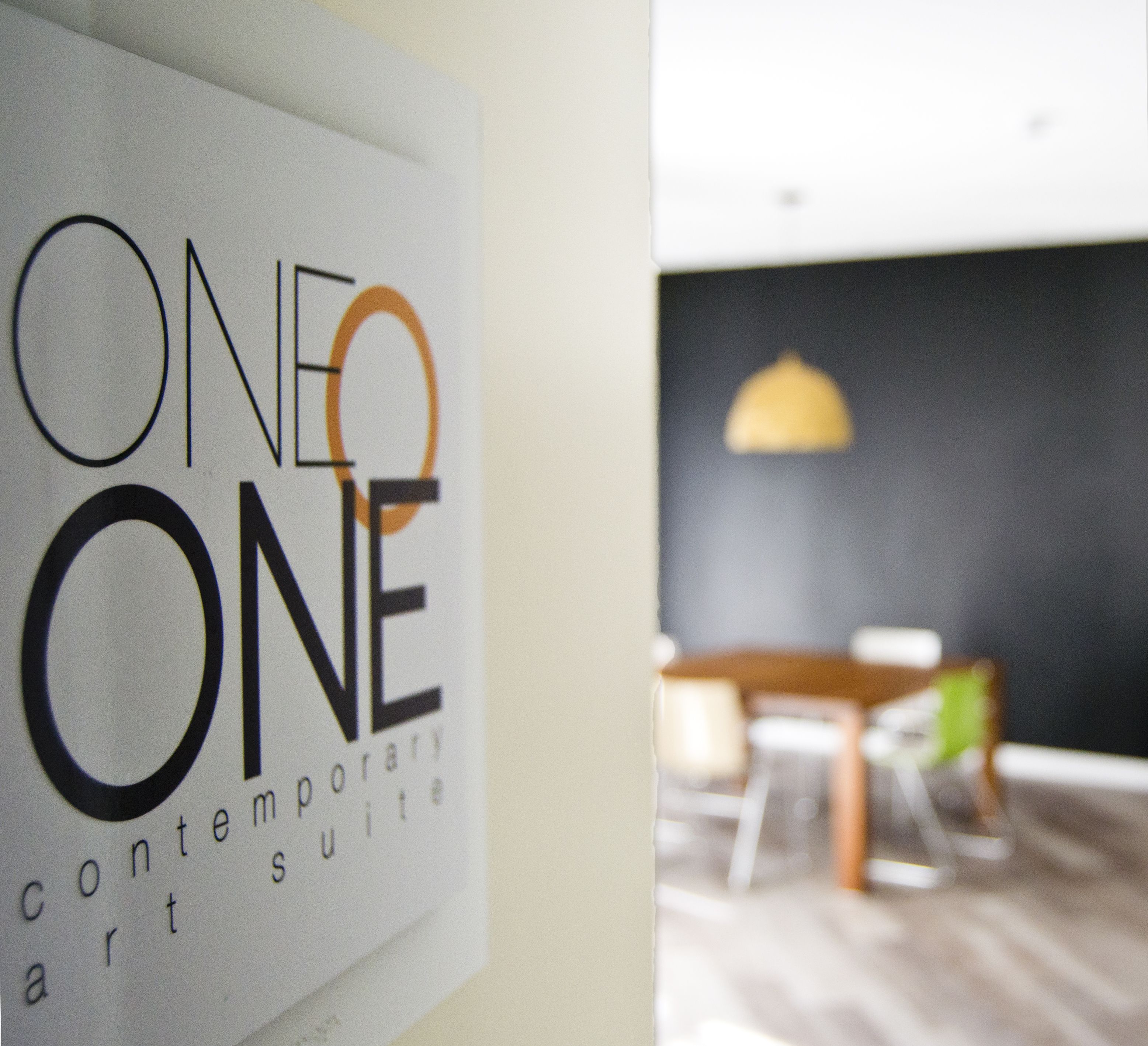 A sign with the text "ONE ONE contemporary art suite" and a blurred interior view of a room with a table and chairs.