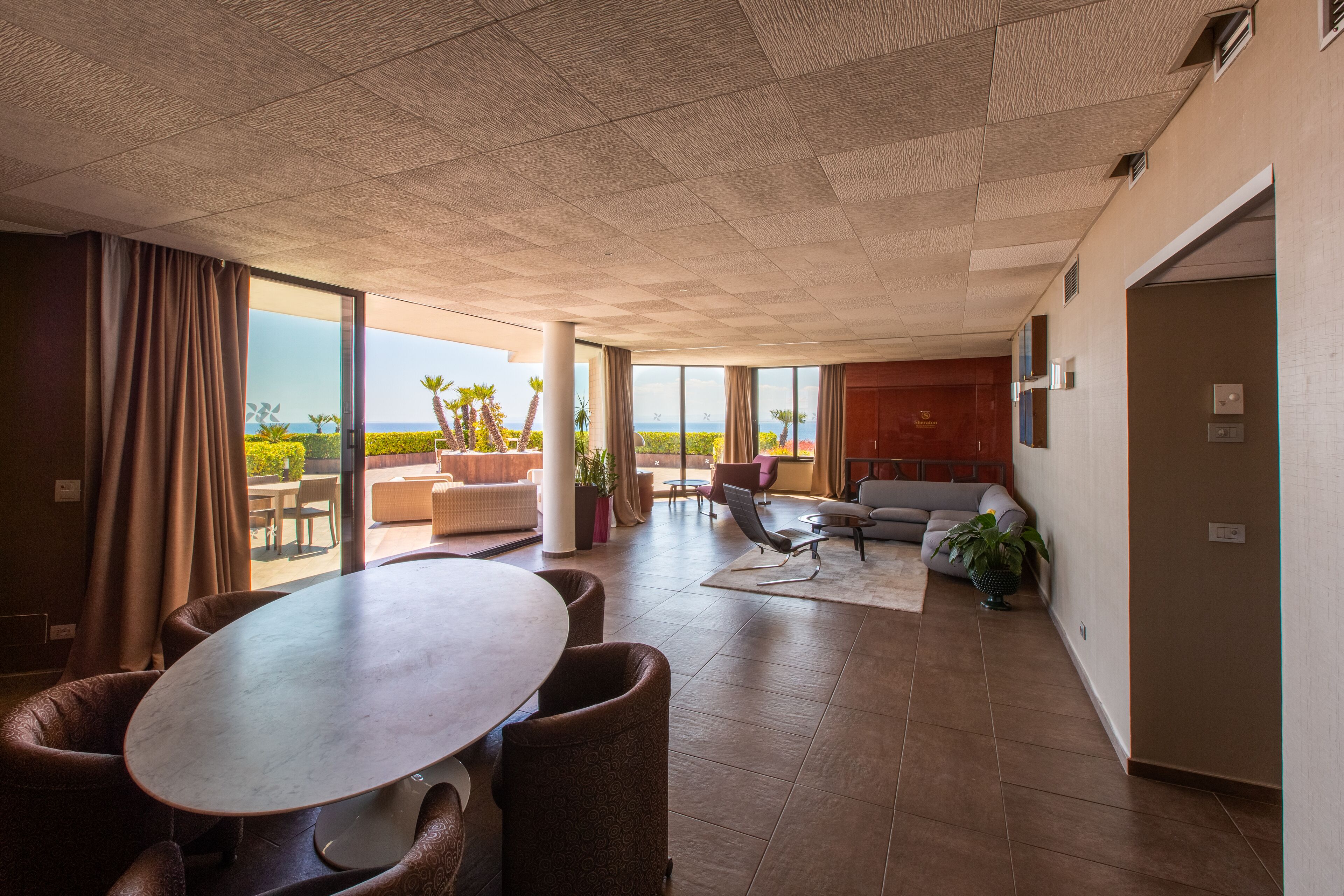 A spacious living area with a large table and comfortable seating, large windows offering a view of the outdoors, and a modern ceiling design.