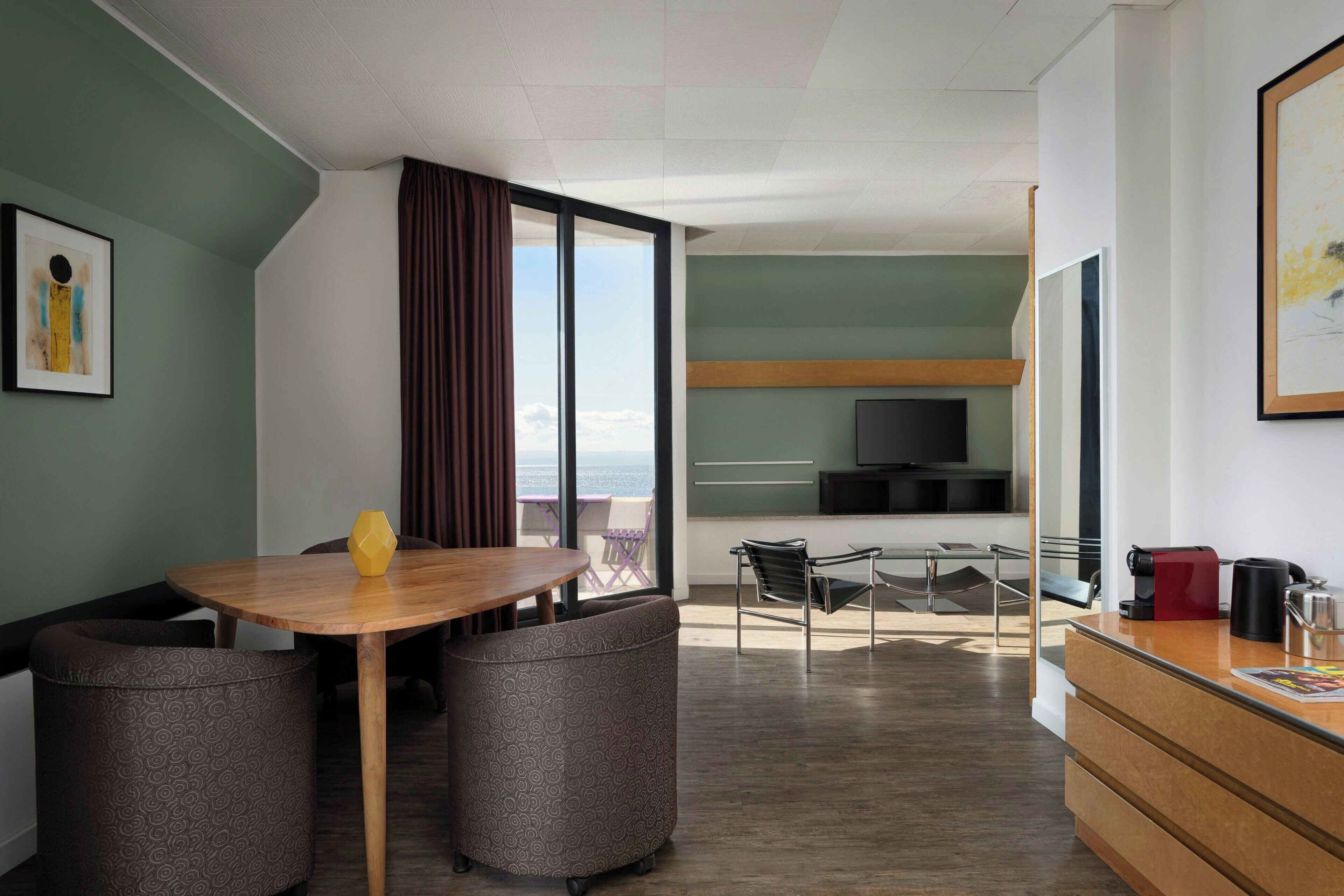 Una moderna camera d'hotel con un tavolo da pranzo in legno, due poltrone a motivi decorativi, un divano, un balcone con vista, una TV e una macchina per il caffè.