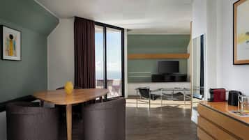 Una moderna camera d'hotel con un tavolo da pranzo in legno, due poltrone a motivi decorativi, un divano, un balcone con vista, una TV e una macchina per il caffè.