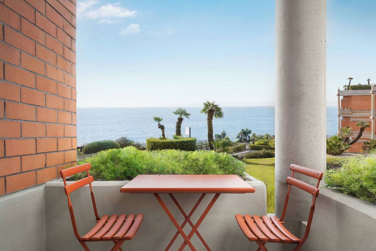 Een balkon met een tafel en twee stoelen, met uitzicht op een aangelegde tuin en de zee.