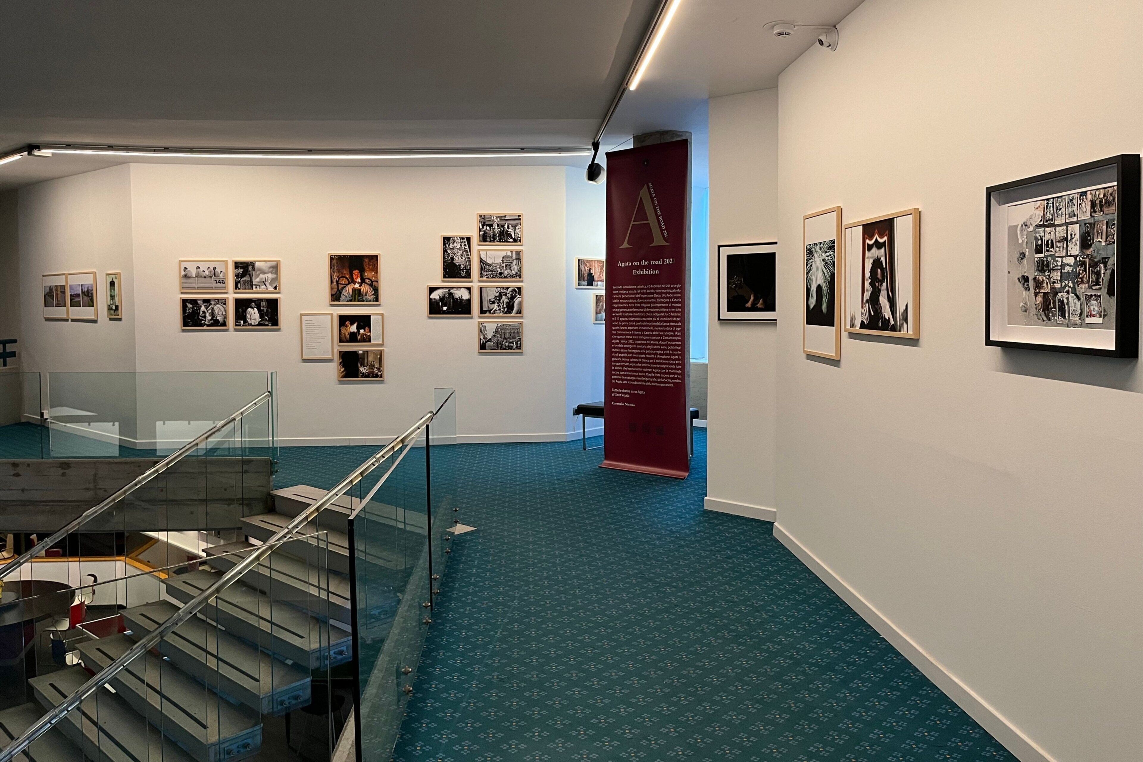 Uma exposição de arte em ambiente interno, com fotografias emolduradas nas paredes, uma escada com guarda-corpos de vidro e um totem informativo vermelho.