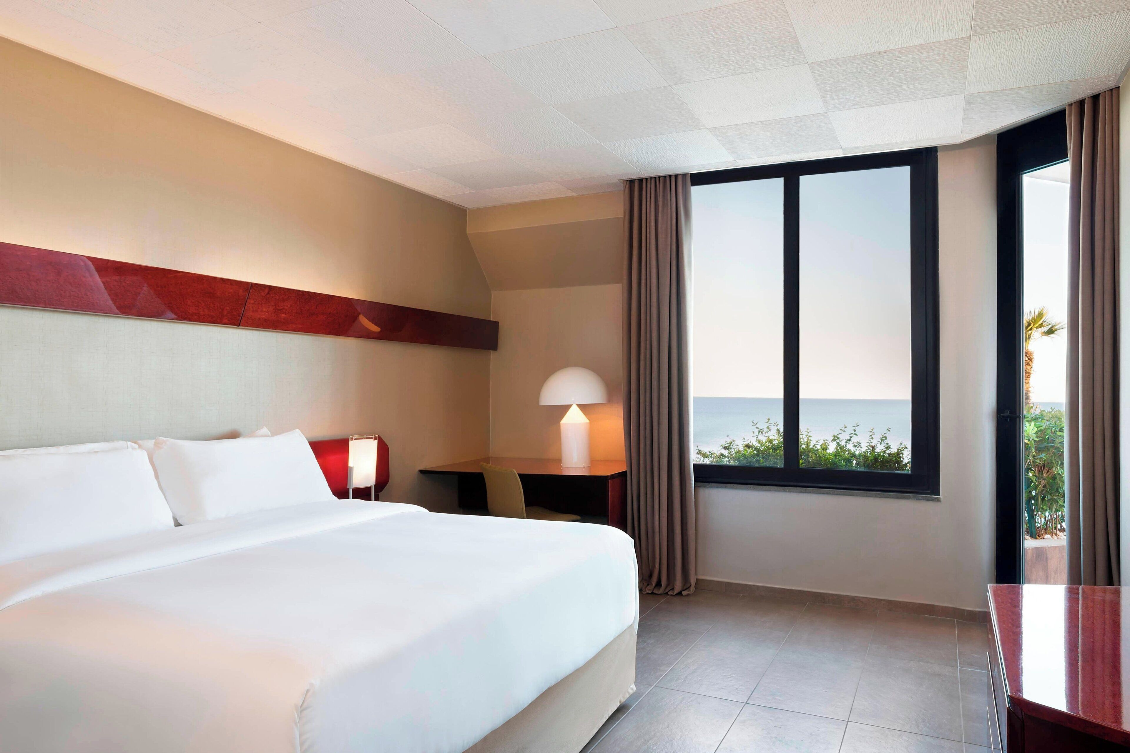 Ein Hotelzimmer mit einem großen Bett, einem Schreibtisch mit Lampe und Blick auf das Meer durch ein Fenster.
