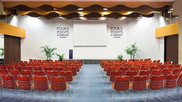 Un salón de conferencias con hileras de sillas anaranjadas, un escenario con una pantalla y dos letreros que dicen "FOUR POINTS BY SHERATON CATANIA".