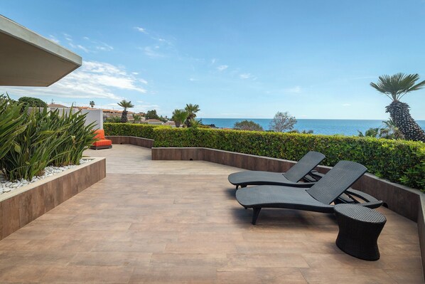 Un patio al aire libre iluminado por el sol con dos sillones, un muro bajo con plantas y vista al mar.