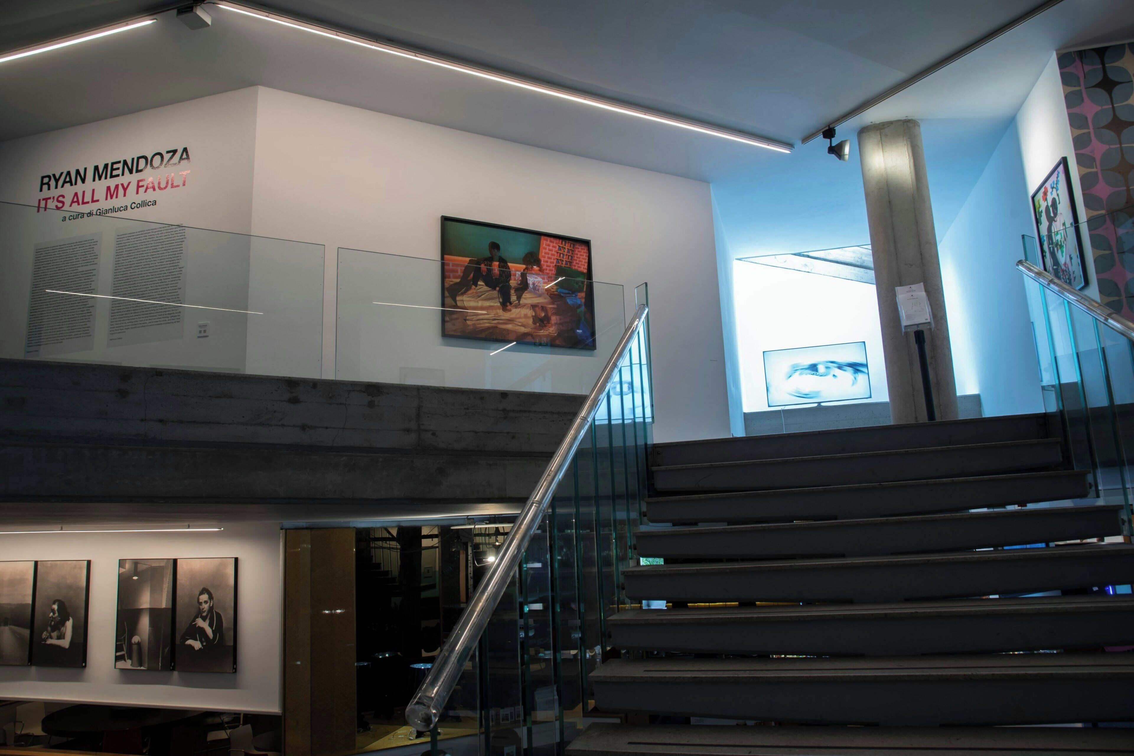 A modern interior with a staircase, artwork on the walls, and a sign for 'Ryan Mendoza - It's All My Fault'.
