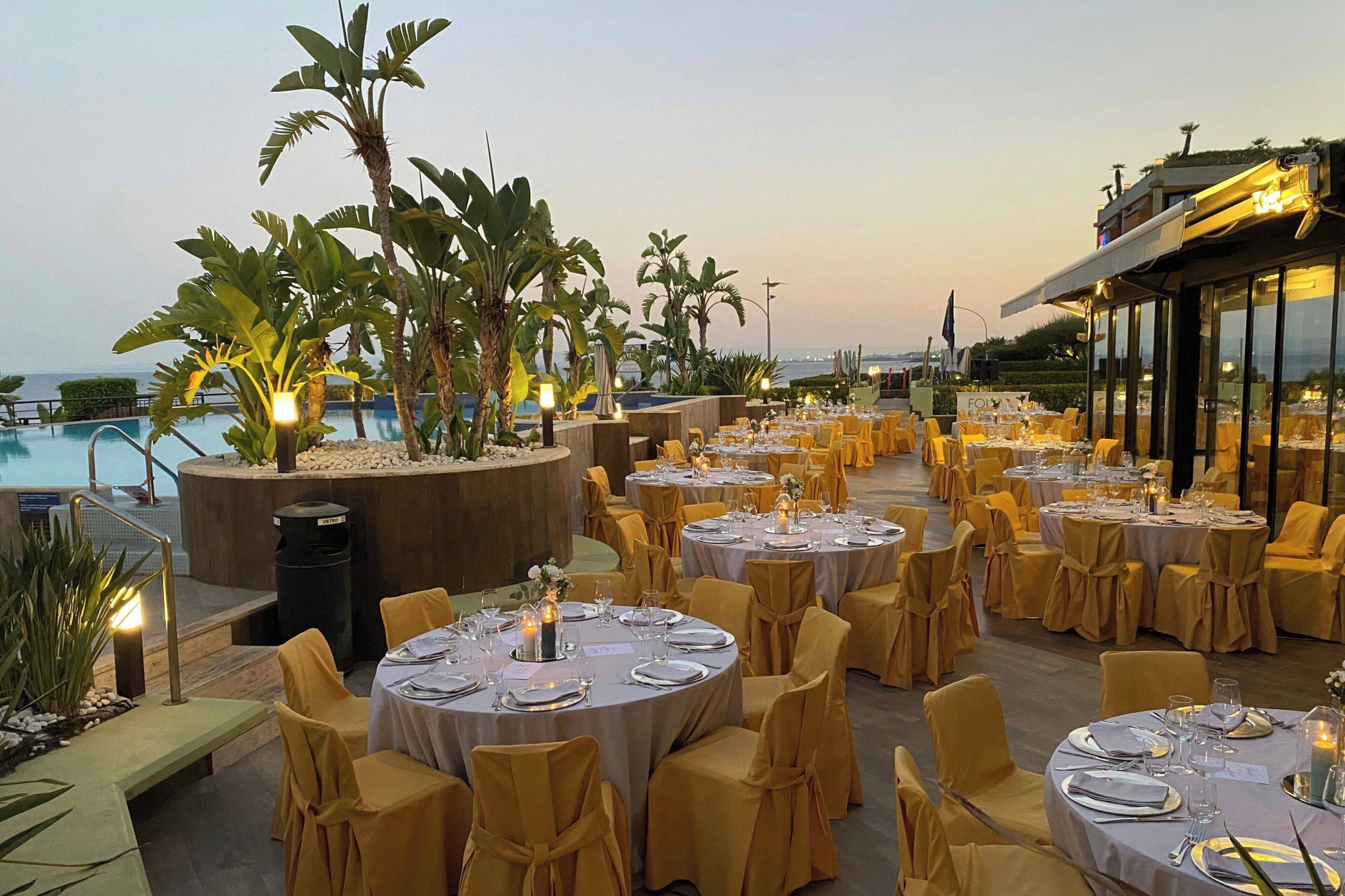 An outdoor dining area with round tables set for a formal event, surrounded by yellow chairs and plants.